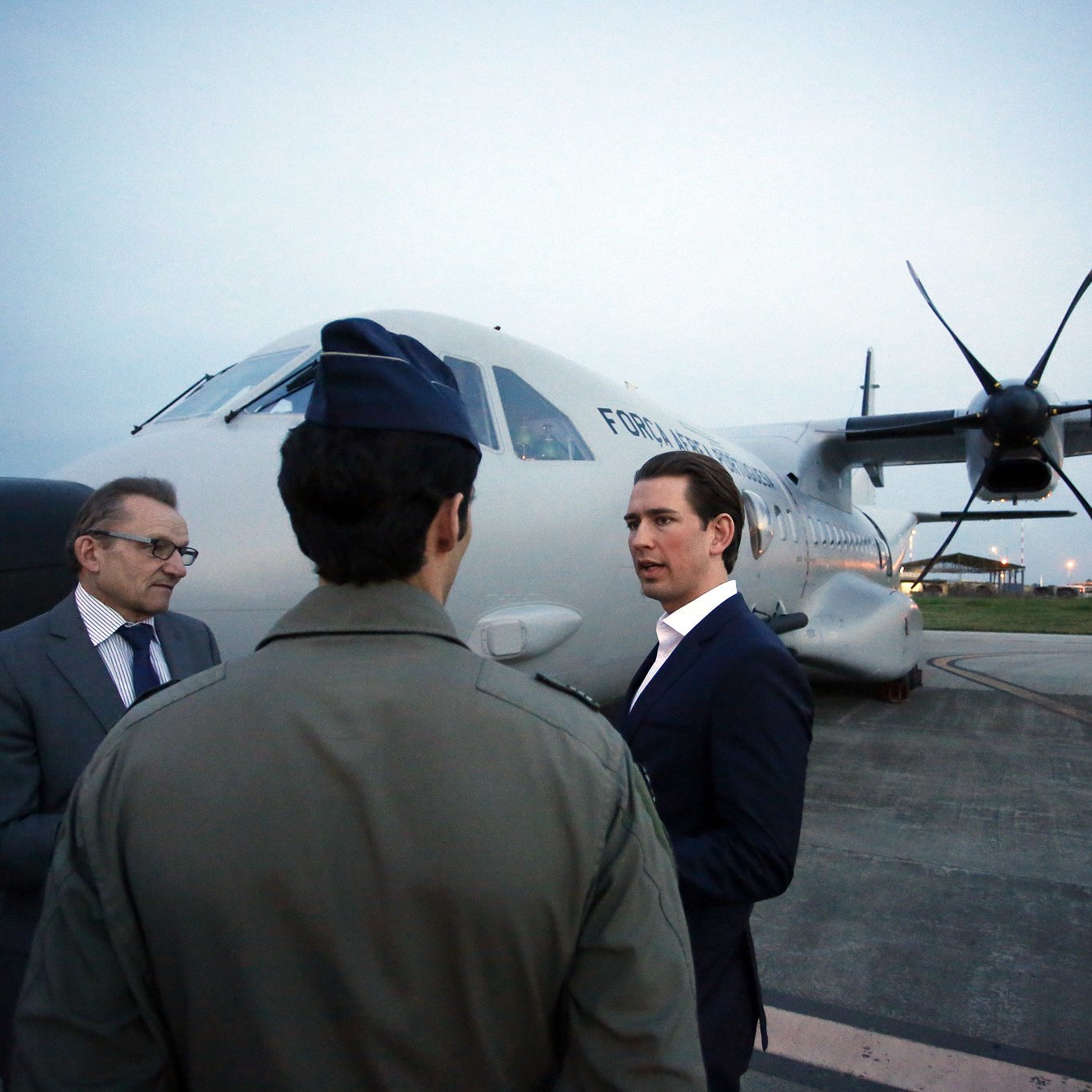 L'image représente un groupe de trois hommes se tenant près d'un avion sur une piste d'atterrissage. L'avion a des hélices et semble être un appareil de taille moyenne. L'éclairage est doux, indiquant qu'il peut être tôt le matin ou tard en soirée. Un homme en costume noir discute avec un autre homme habillé d'une tenue militaire. Le troisième homme, portant des lunettes, semble écouter la conversation. L'environnement est urbain et l'aéroport est visible en arrière-plan avec quelques lumières. L'atmosphère est professionnelle et engageante.