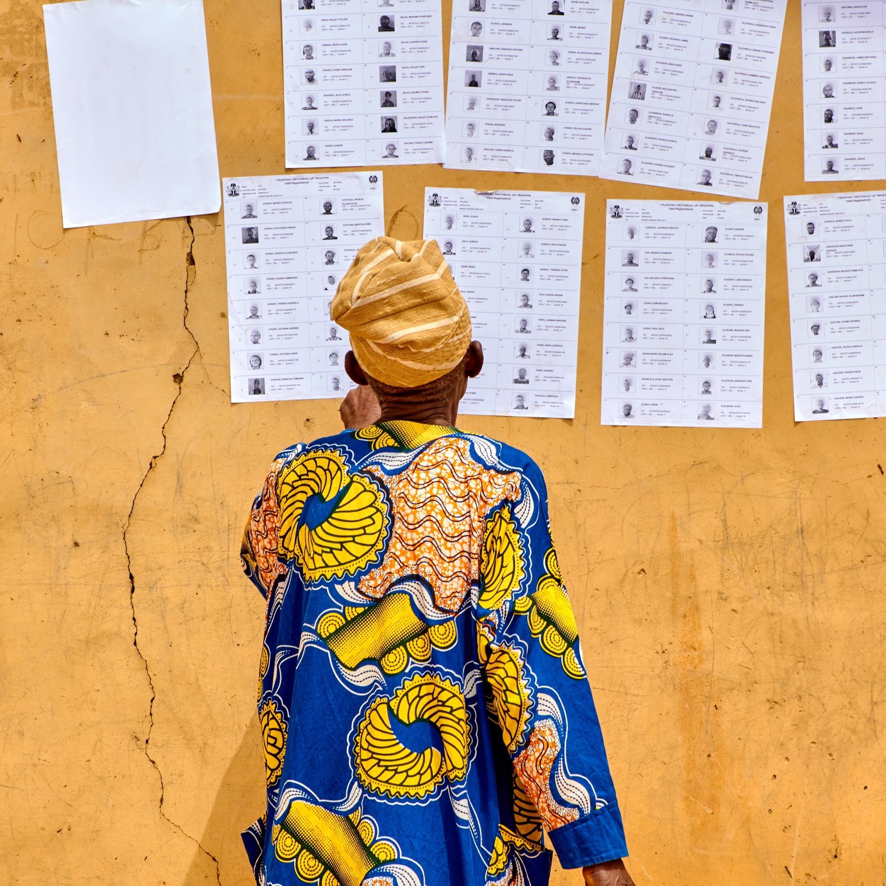 L'image montre un homme de dos, vêtu d'une robe colorée avec des motifs traditionnels, principalement en bleu et jaune. Il se tient devant un mur de couleur ocre, où sont affichés plusieurs documents. Ces documents ressemblent à des bulletins de vote ou des listes, alignés verticalement, avec des photographies et des noms inscrits. Le sol est légèrement fissuré, ajoutant une texture au fond de l'image. L'homme porte un turban beige, et son attention semble focalisée sur les informations affichées sur le mur.
