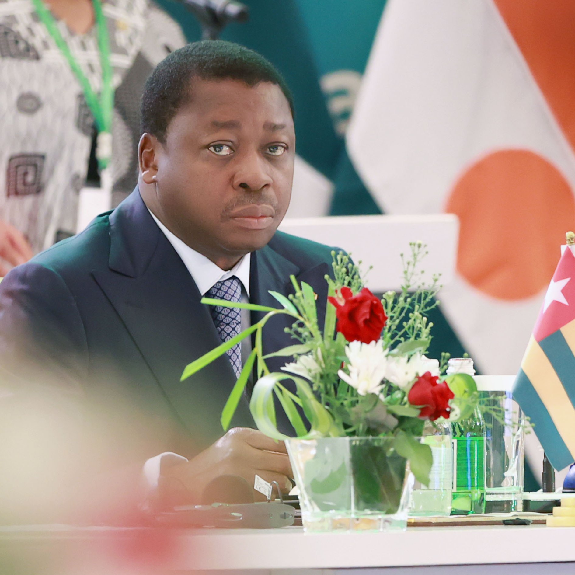 L'image montre un homme assis à une table lors d'un événement officiel. Il porte un costume sombre et une cravate, et son expression semble sérieuse et attentive. Sur la table, il y a un petit vase avec des fleurs colorées, ajoutant une touche de vie à la scène. À côté de lui, on voit un drapeau qui représente le Togo. En arrière-plan, des drapeaux d'autres pays sont également visibles, indiquant un cadre international. L'atmosphère semble formelle et concentrée.