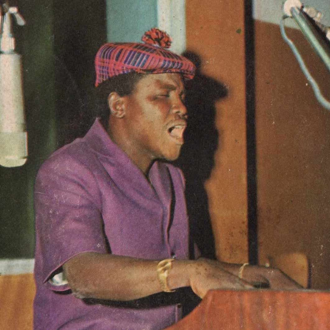 Sur cette image, nous voyons un homme en train de jouer du piano. Il porte une chemise violette et un chapeau à motif écossais. Son expression est concentrée et passionnée alors qu'il chante. Deux microphones sont placés à proximité, capturant sa voix. L'arrière-plan présente des panneaux en bois colorés, créant une ambiance chaleureuse et artistique. L'atmosphère dégage une sensation de musique vivante et d'émotion.