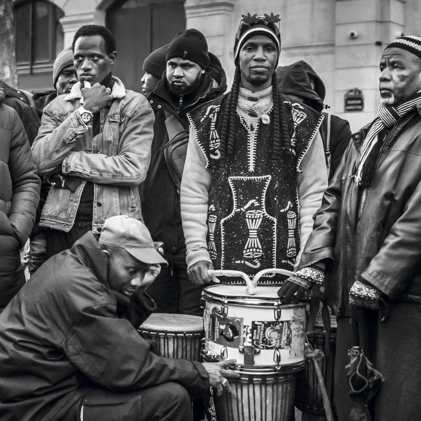 L'image montre un groupe de plusieurs hommes rassemblés dans un environnement urbain. Ils portent des vêtements variés, allant de parkas sombres à des vêtements traditionnels. Certains d'entre eux regardent dans des directions différentes, affichant une diversité d'expressions. Au centre, un homme est assis, jouant sur un tambour, ajoutant une ambiance musicale à la scène. Les autres semblent attentifs, créant une atmosphère de camaraderie et d'échange. L'image est en noir et blanc, accentuant les contrastes et les textures des vêtements et des visages.