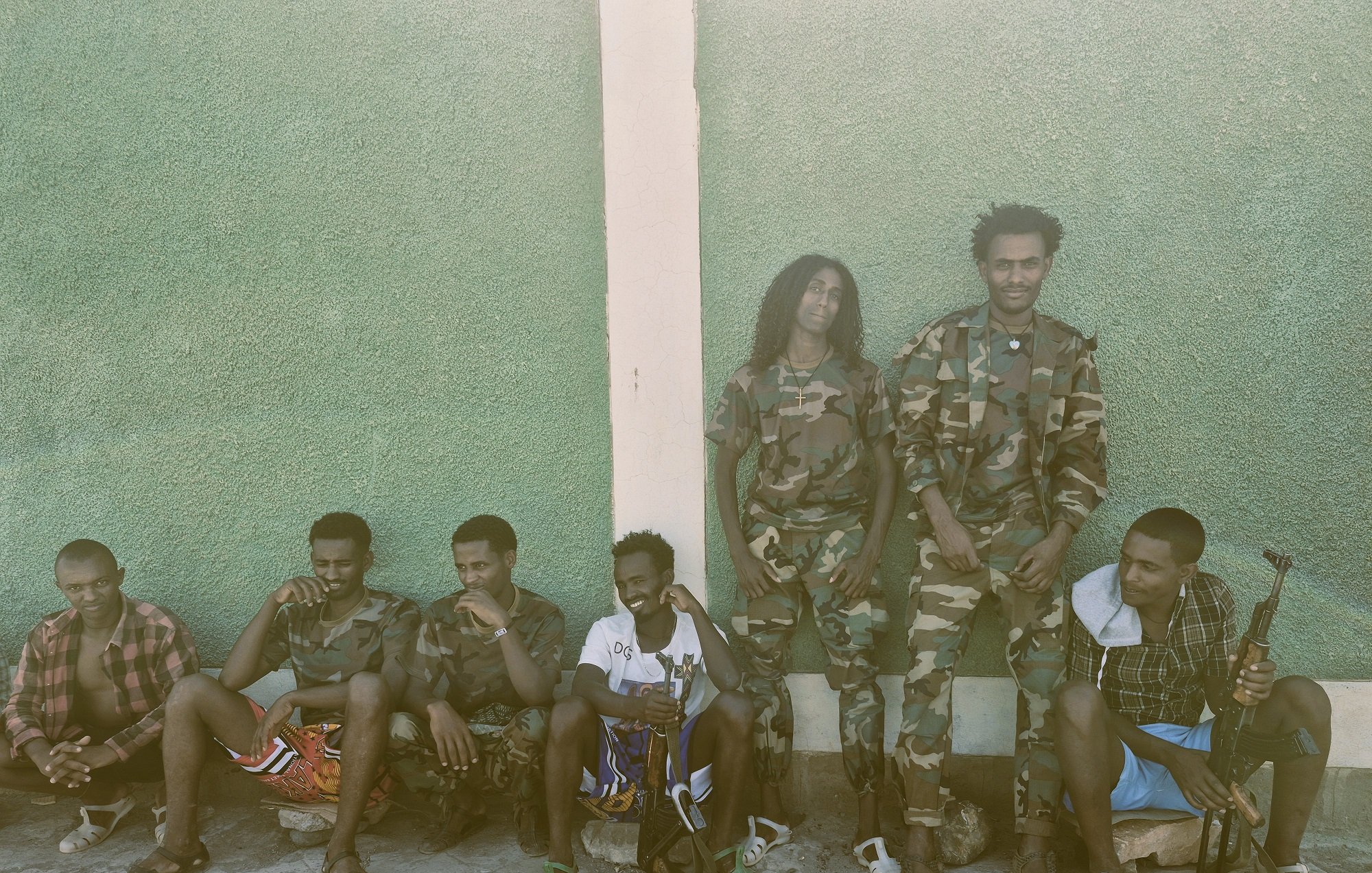 L'image montre un groupe de jeunes hommes, assis et debout, le long d'un mur vert. Les hommes portent des uniformes militaires camouflés pour certains, et d'autres sont habillés de manière plus décontractée. Ils semblent être dans un environnement extérieur, avec un sol en pierre. Quelques-unes des personnes tiennent des objets qui ressemblent à des sacs ou des équipements militaires. L'ambiance est tranquille, mais dégage une impression de camaraderie parmi le groupe.