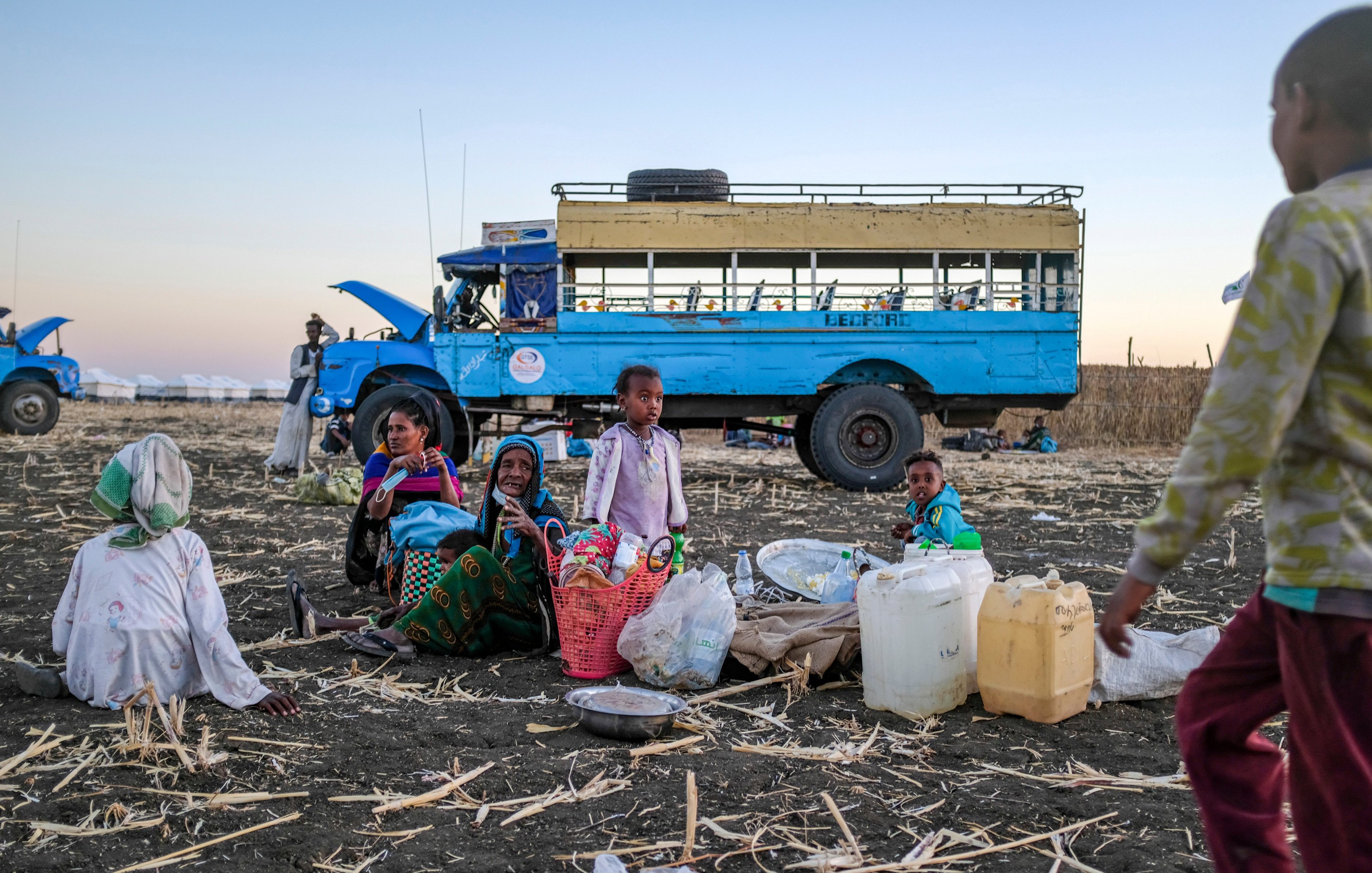 L'image montre un groupe de personnes assises sur un sol de terre battue, entourées de paniers et de contenants. Au centre, des femmes et des enfants semblent converser, tandis qu'un enfant se tient à droite, s'approchant. En arrière-plan, on peut voir un bus bleu immobile, avec un ciel dégradé qui passe des teintes orangées aux bleues, suggérant un crépuscule. Le terrain est jonché de restes de culture, ajoutant un contexte rural à la scène. L'atmosphère évoque une ambiance de rassemblement communautaire dans un environnement de travail.