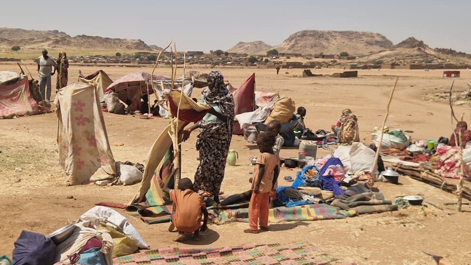 L'image montre un campement dans une région aride et désertique. On peut imaginer une vaste étendue de terrain sec, avec quelques collines en arrière-plan. Des tentes rudimentaires, faites de toile ou de vêtements usagés, sont disposées de manière désordonnée, offrant un semblant de refuge aux personnes qui y vivent. Au sol, des tapis ou des morceaux de tissu sont étalés, tandis que des objets personnels, comme des sacs et des casseroles, sont éparpillés autour. Au centre de la scène, un groupe de personnes est en train de s'activer : une femme semble organiser quelque chose, tandis qu'un enfant debout à côté d'elle observe. L'atmosphère transpire la précarité mais aussi une certaine résilience face aux conditions difficiles. Les couleurs des tissus sont variées, mais plutôt ternes et fanées par le temps. L'ensemble dégage une impression de lutte pour la survie dans un environnement hostile.