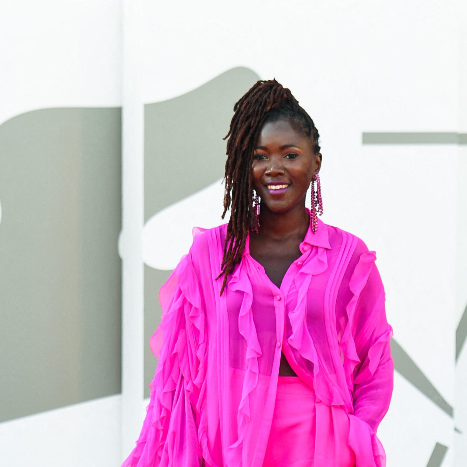 L'image montre une femme debout sur un fond minimaliste. Elle porte une tenue éclatante de couleur rose vif, composée d'une blouse légère aux manches froncées et d'une jupe assortie. Son sourire est radieux et elle affichent une attitude pleine de confiance. Ses cheveux sont coiffés en dreadlocks, et elle porte de grandes boucles d'oreilles qui ajoutent une touche élégante à son look. Le fond est composé de formes géométriques en tons neutres, mettant en valeur la vivacité de sa tenue.