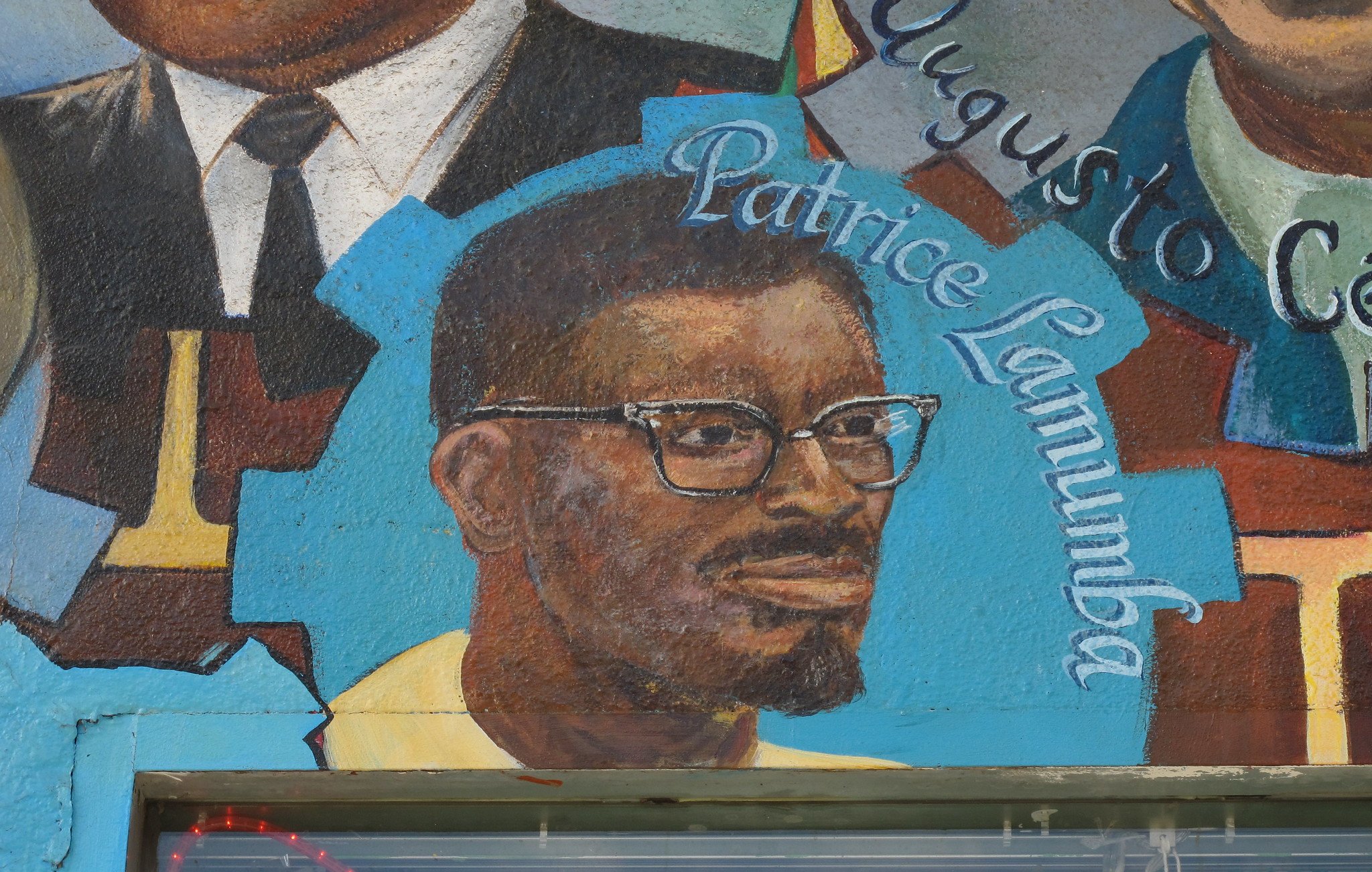 L'image semble représenter une fresque murale avec des portraits de différentes personnes. Au centre, on peut voir un homme portant des lunettes, avec une barbe et un sourire. Son nom, "Patrice Lumumba", est écrit en dessous de son portrait. Les autres éléments de la fresque peuvent inclure des symboles ou des motifs colorés, contribuant à une ambiance artistique et culturelle. La fresque pourrait célébrer des figures historiques ou politiques importantes.