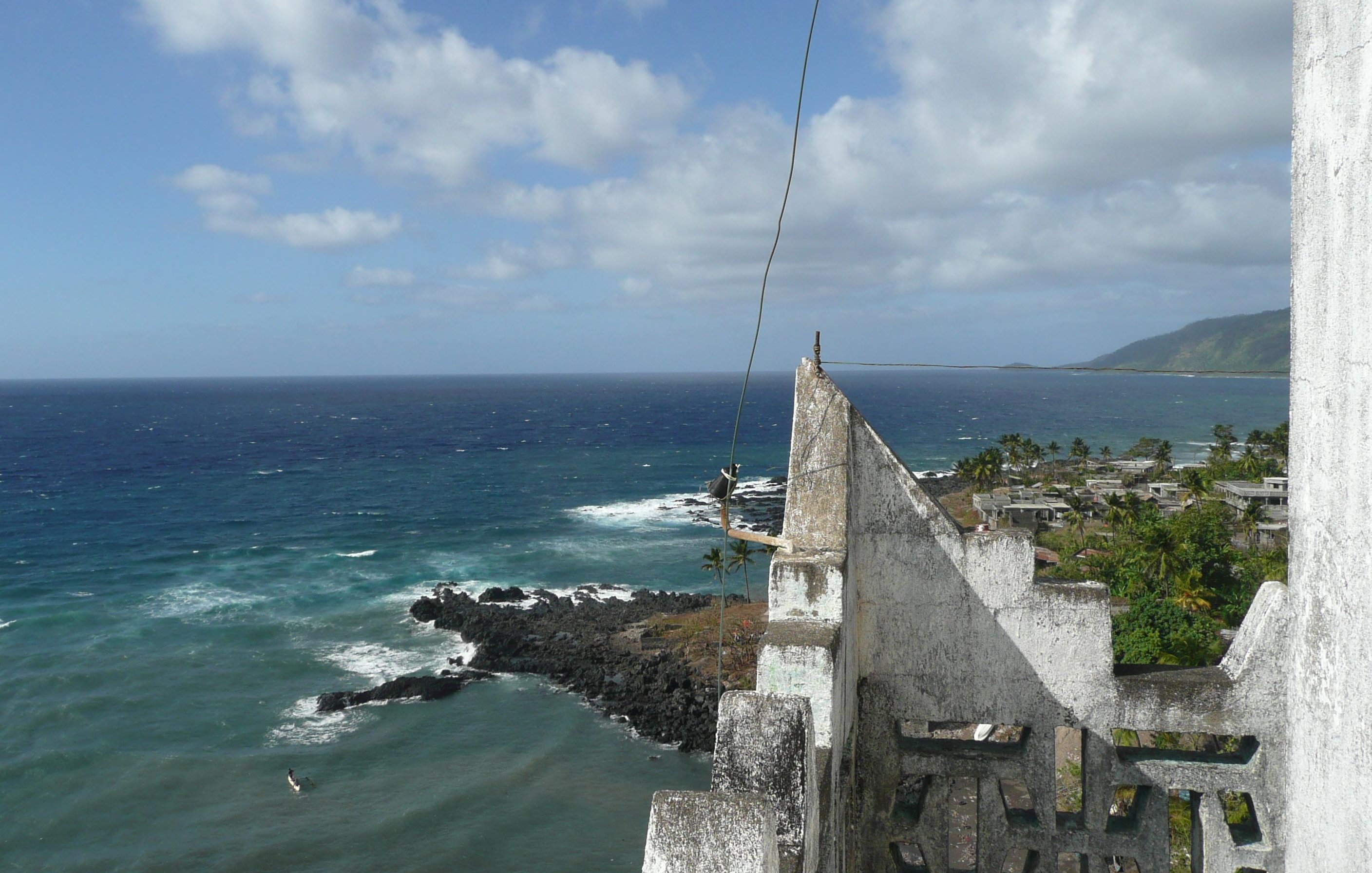 Imaginez-vous au sommet d'une vieille tour, où l'air est frais et légèrement salé. Devant vous s'étend un vaste océan d'un bleu profond, parsemé de vagues qui dansent et s'écrasent sur des rochers sombres. Au loin, vous pouvez percevoir la côtes, avec des palmiers se balançant doucement au gré du vent. Le ciel est parsemé de nuages blancs qui flottent, créant un jeu de lumière sur l'eau. Sur votre droite, des paysages verdoyants de collines se dessinent, ajoutant à la beauté sauvage de cet endroit. C'est un panorama paisible et apaisant, où la nature semble en harmonie avec l'horizon.