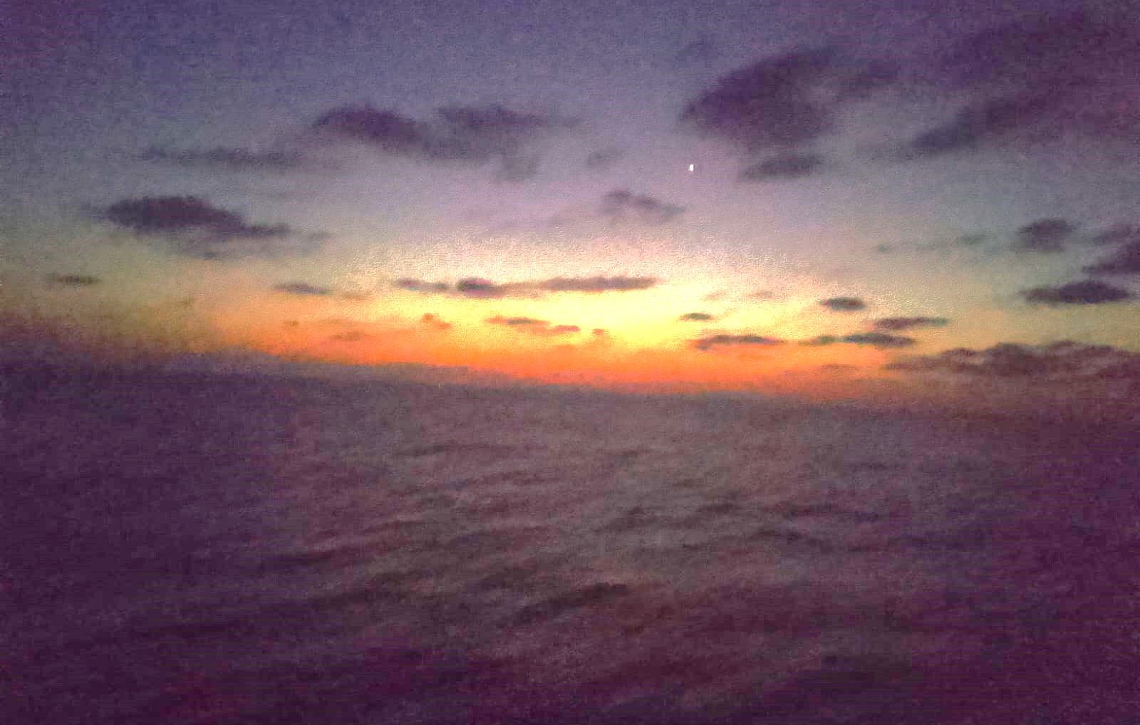 Cette image montre un paysage maritime au crépuscule. Le ciel est teinté de nuances sombres, avec des nuages épars. On remarque un doux dégradé de couleurs allant du bleu foncé au orange, signalant la transition entre le jour et la nuit. L'horizon est légèrement flou, et l'eau semble calme, reflétant les couleurs du ciel. Une étoile brillante est visible dans la partie supérieure de l'image, ajoutant une touche de mystère à cette scène sereine et nocturne. L'atmosphère générale est paisible et contemplative.