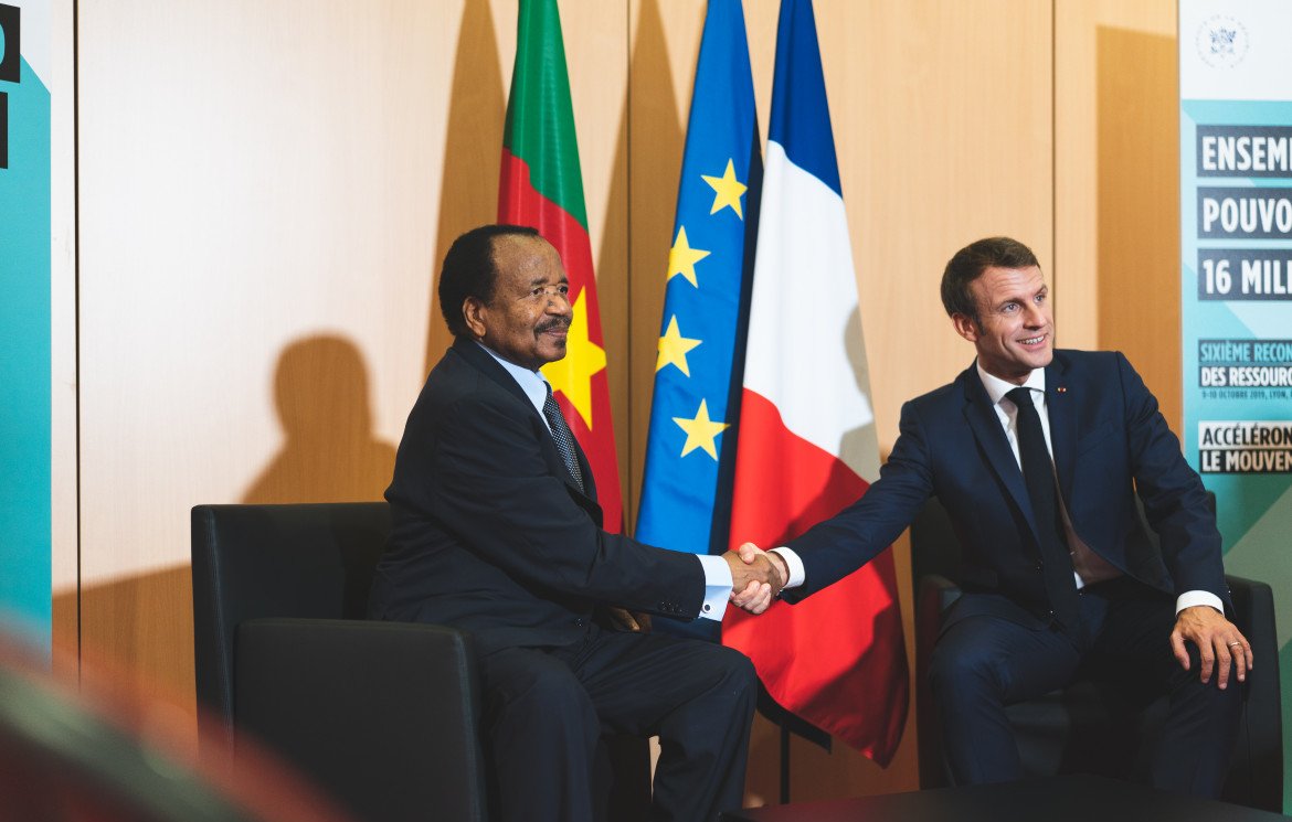 Dans cette image, deux hommes sont assis l'un en face de l'autre, se serrant la main dans un geste formel. L'homme à gauche porte un costume sombre et a une expression souriante. À sa droite, un autre homme, également en costume, affiche un sourire poli. En arrière-plan, on peut noter deux drapeaux : l'un est le drapeau du Cameroun, l'autre le drapeau français et le drapeau de l'Union européenne. L'environnement semble être un espace de réunion officiel, avec des panneaux informatifs visibles. L'atmosphère dégage une impression de cordialité et de coopération.