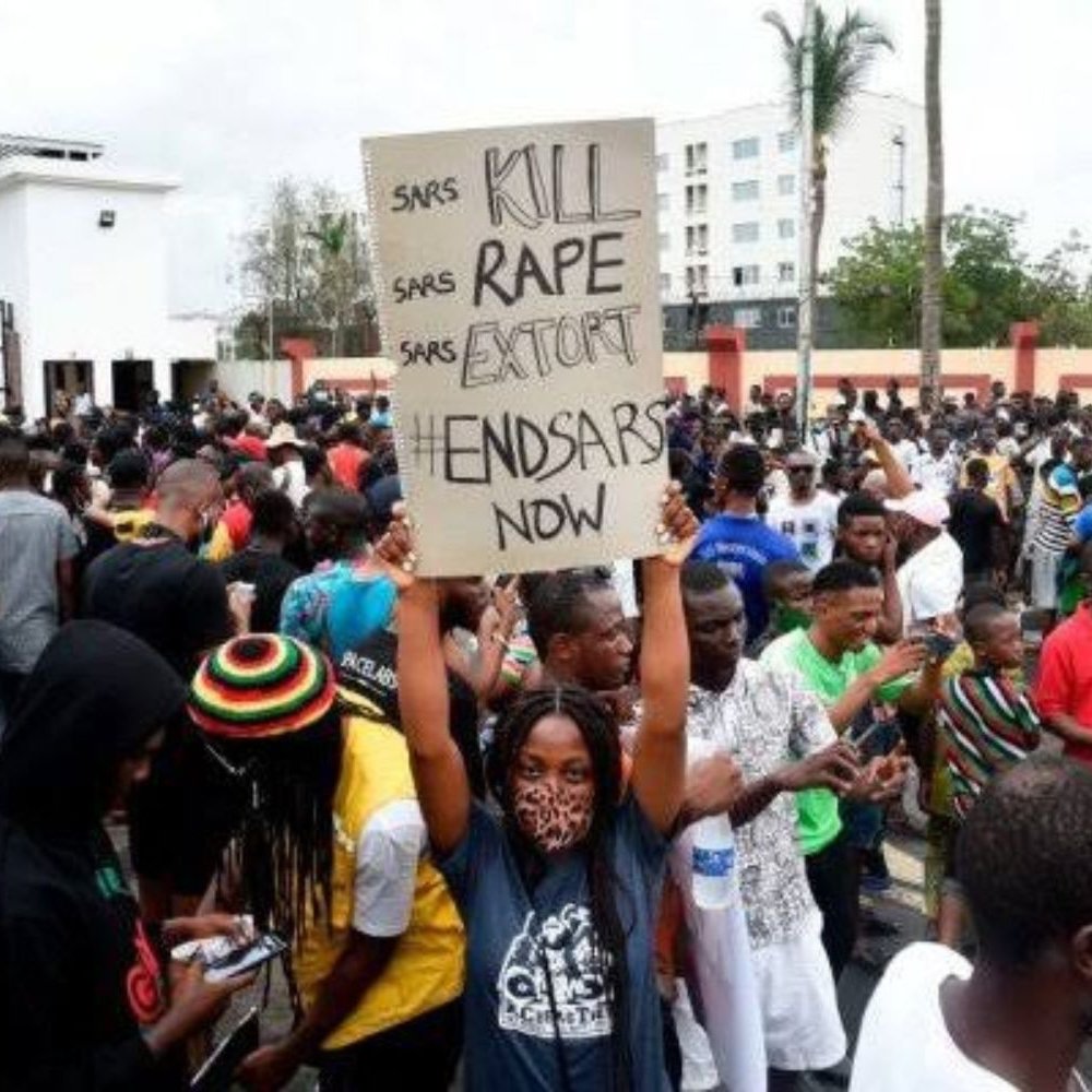 L'image montre une foule dense rassemblée pour une manifestation. Au centre, une personne tient une grande pancarte avec des inscriptions demandant la fin d'abus de la police. Les manifestants portent des vêtements variés, certains affichent des signes de solidarité, comme des masques. En arrière-plan, on aperçoit des bâtiments et des palmiers, ce qui donne une idée que l'événement se déroule dans une ville. L'atmosphère est chargée d'énergie collective, marquée par un sentiment de lutte et de revendication pour des droits.