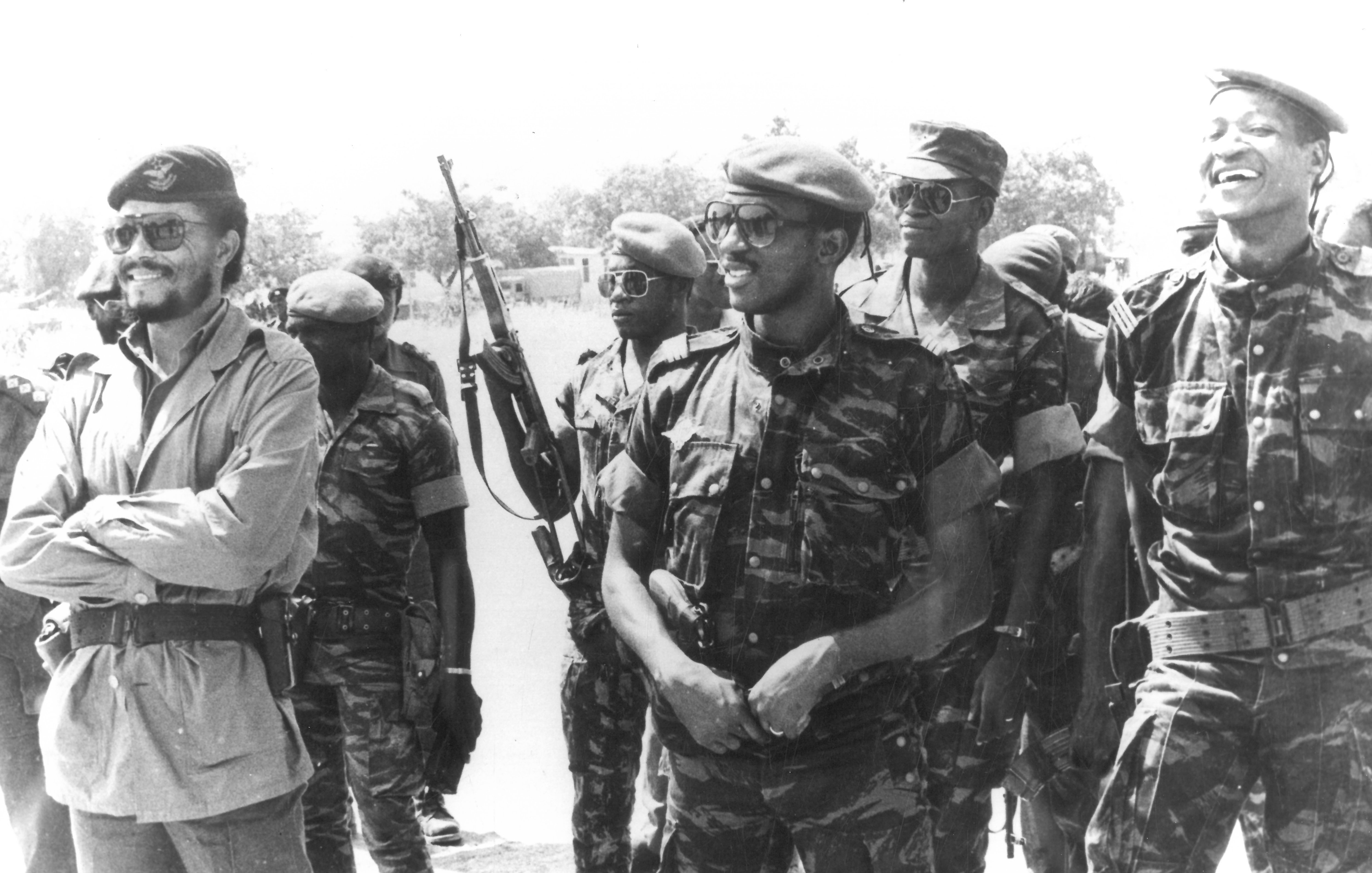 Cette image en noir et blanc montre un groupe de soldats, tous habillés en uniforme militaire. Au centre, un homme souriant porte un beret et un uniforme camouflage. À sa droite, un autre homme, également en uniforme, affiche un large sourire. En arrière-plan, plusieurs soldats tiennent des fusils et portent des lunettes de soleil. L'ambiance semble détendue, avec des expressions de camaraderie parmi les membres du groupe. Le décor en arrière-plan est peu détaillé, mais on peut deviner un environnement extérieur.