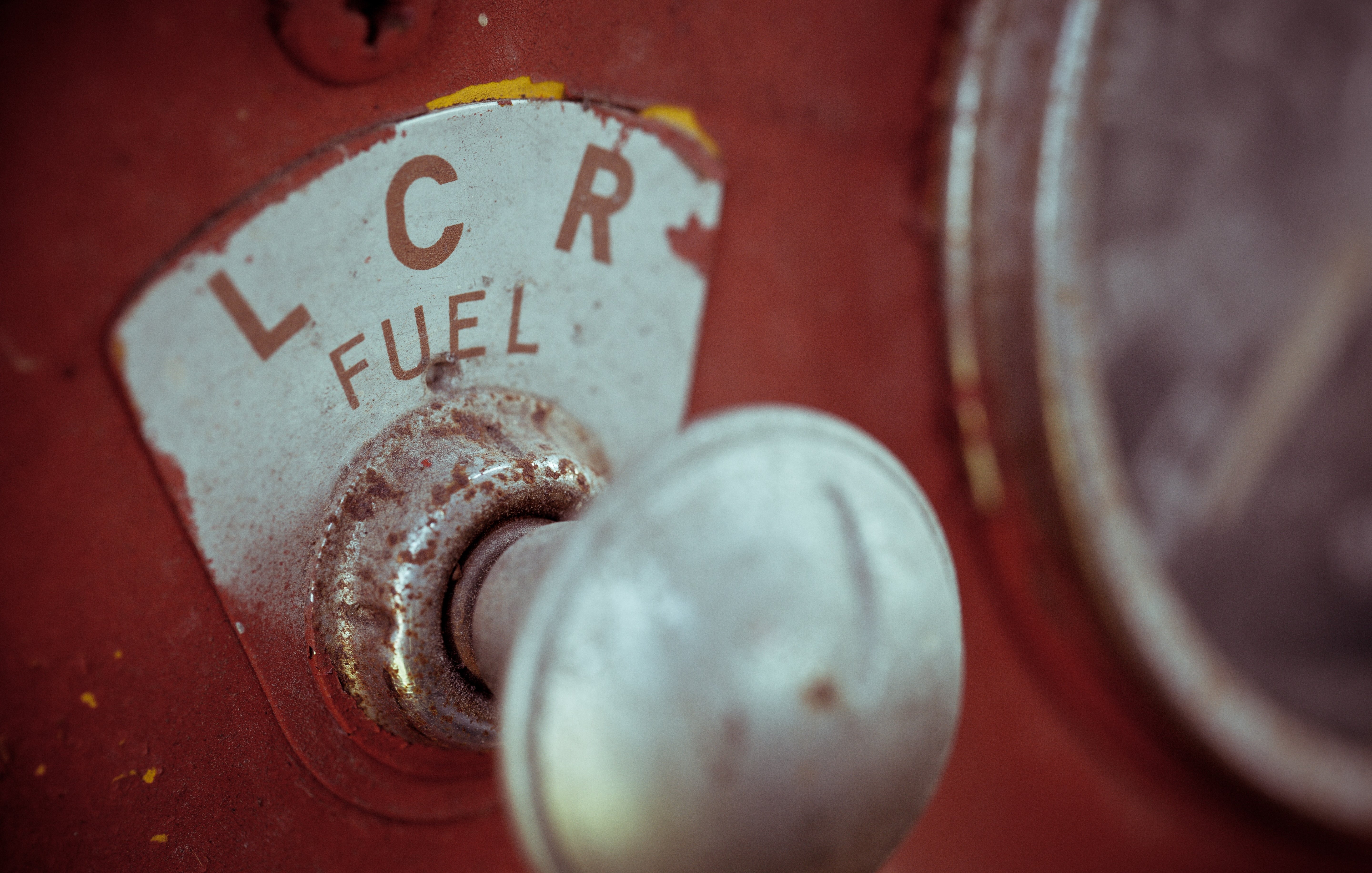 L'image montre un gros plan d'un ancien indicateur de carburant sur une machine, probablement un tracteur ou un véhicule vintage. La partie visible présente un disque circulaire marqué avec les lettres "LCR" et le mot "FUEL" en dessous. La surface est usée, avec des éraflures et une couleur rouge qui témoigne de son âge. À côté, il y a un bouton en métal brillant, légèrement terni, qui ajoute à l'aspect nostalgique de l'objet. L'ensemble évoque une impression de robustesse et d'histoire, typique des équipements anciens.