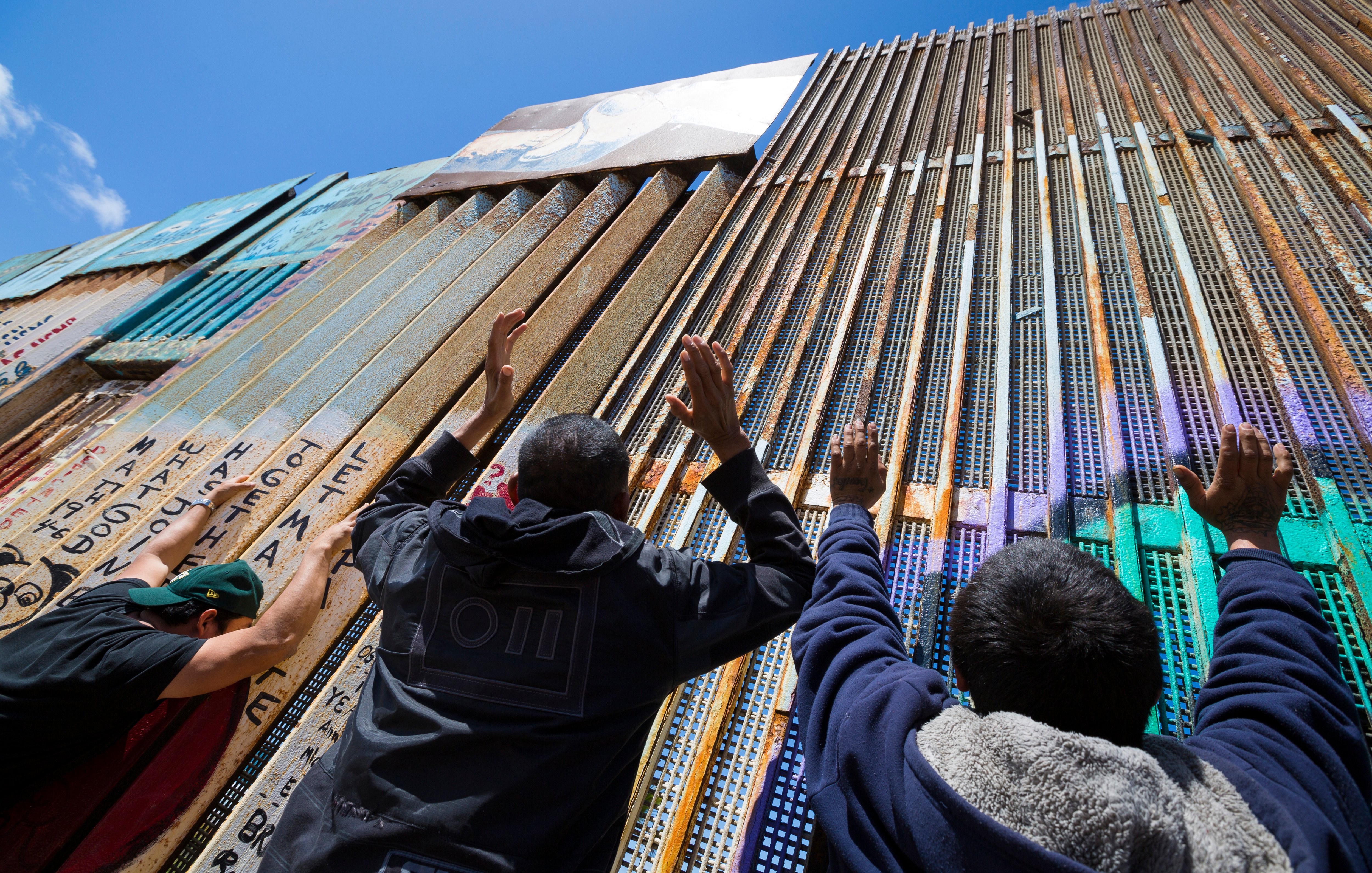 Sur cette image, on peut voir un grand mur en métal qui sépare deux zones. Le mur est composé de panneaux de différentes textures et couleurs, certains recouverts de graffitis. Au premier plan, on aperçoit plusieurs personnes qui tendent les mains vers le mur, comme si elles cherchaient un contact ou un lien. Le ciel est bleu et dégagé en arrière-plan, ce qui crée un contraste avec le métal de la barrière. L'atmosphère semble chargée d'émotion et de désir de connexion.