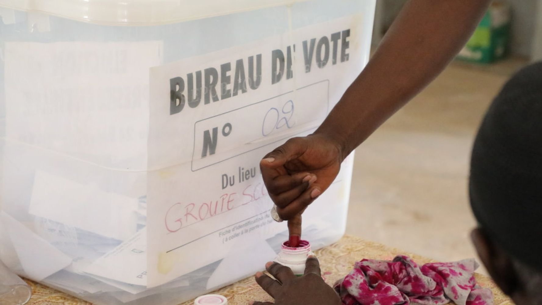 Devant vous se trouve une scène typique d'un bureau de vote. Il y a une urne transparente avec une étiquette indiquant "BUREAU DE VOTE N° 02" et le lieu sur une feuille, probablement fixée à l'urne. Un électeur est en train de voter : sa main est penchée vers une petite boîte de vote où il semble plonger son doigt dans de l'encre rouge. Sur la table, il y a des morceaux de tissu, probablement utilisés pour marquer les bulletins ou pour l'organisation du vote. L'ambiance est calme, avec un sentiment de sérieux et d'engagement civique.