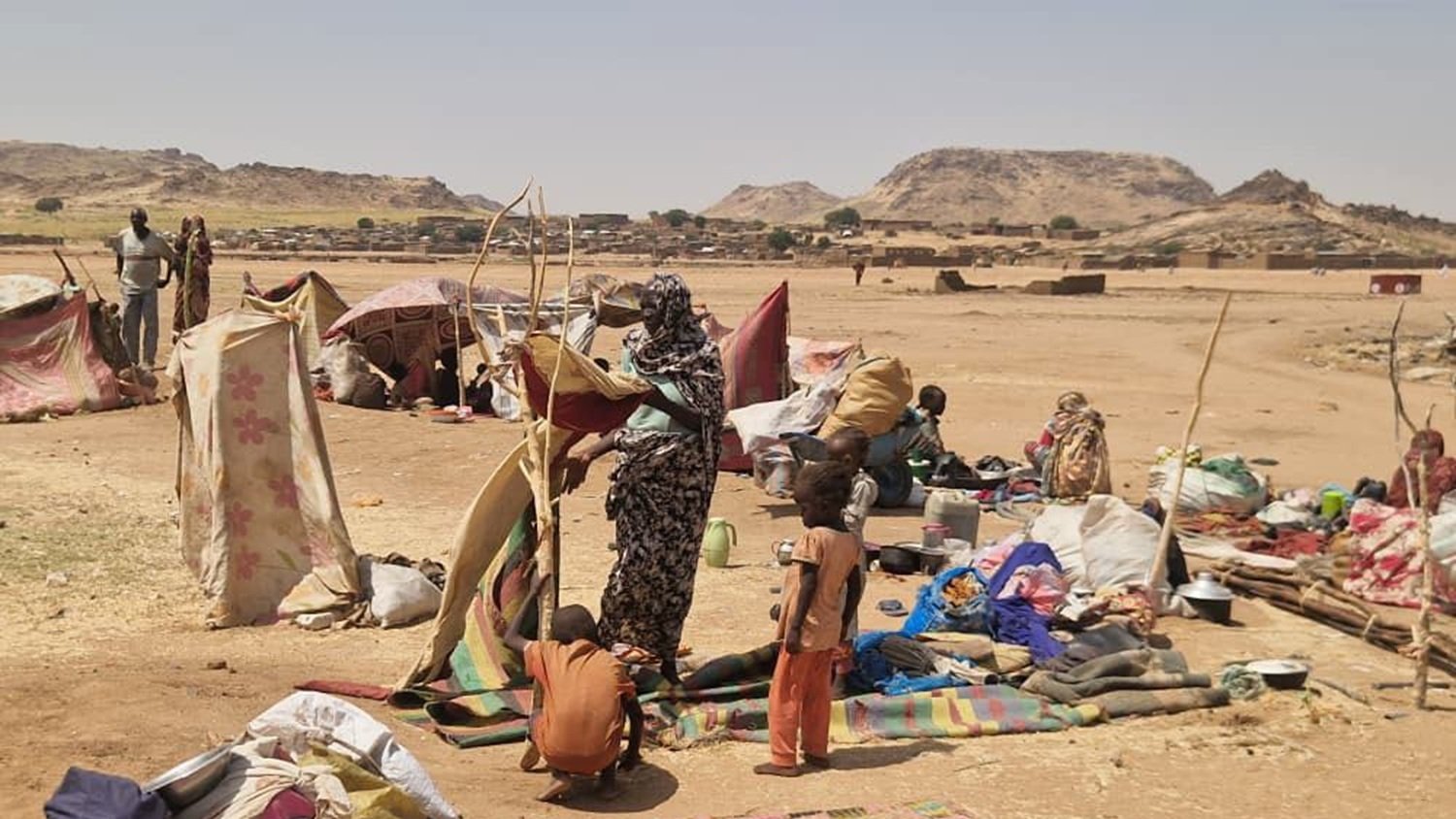 The image depicts a temporary settlement, likely a displaced persons camp or informal shelter in a rugged landscape. Several makeshift tents and tarps are set up, providing shelter for the residents. In the foreground, a woman is seen holding a structure, possibly a pole or support for one of the tents, while a child stands nearby. The ground is scattered with blankets, bags, and various belongings, suggesting a living area that is communal and somewhat crowded. The background shows a barren landscape with hills or rocky formations, indicating a harsh environment.