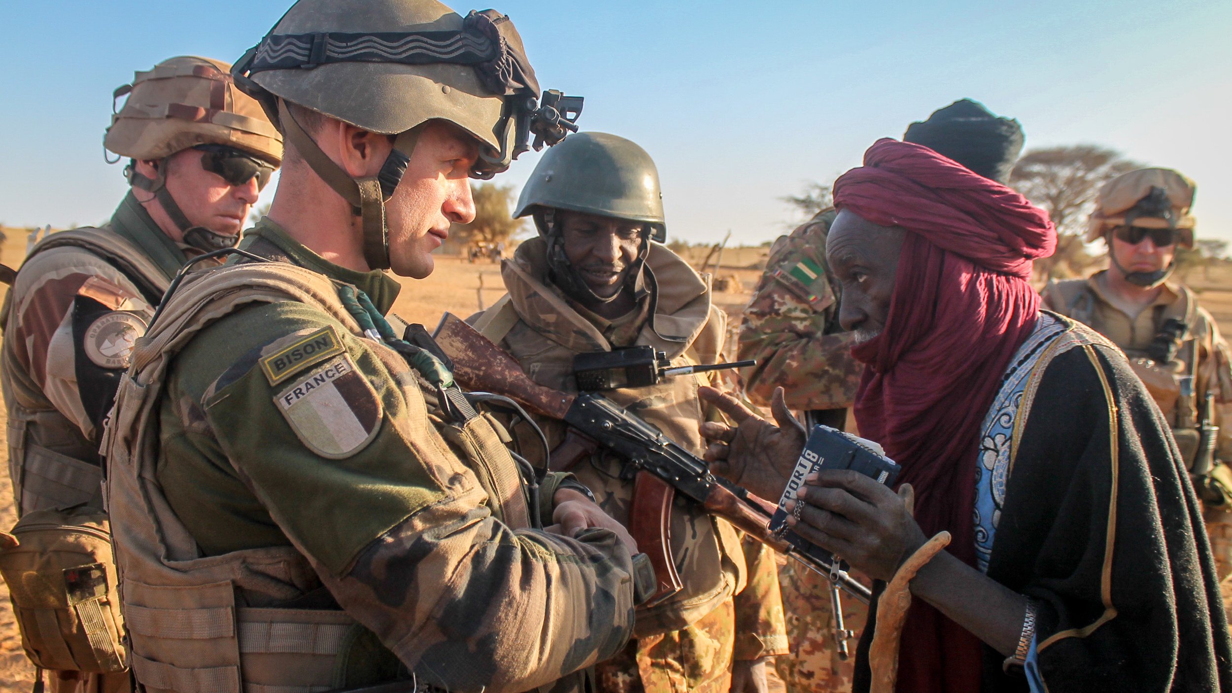 L'image montre une scène dans un environnement désertique, probablement au Sahel. Au premier plan, un soldat en uniforme militaire, portant un gilet pare-balles et un casque, interagit avec une femme vêtue d'une robe traditionnelle. Elle a l'air âgée et semble lui parler tout en tenant un objet dans ses mains. D'autres soldats en arrière-plan, arborant également des uniformes militaires, observent la scène avec un air attentif. L'éclairage est naturel, suggérant que la photo a été prise en fin d'après-midi, et le paysage environnant est sec avec des dunes de sable visibles. Cette rencontre reflète peut-être une interaction humanitaire ou de soutien entre les militaires et la population locale.