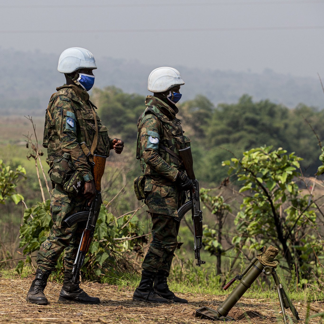 L'image montre deux militaires en uniforme, portant des casques blancs et des masques. Ils se tiennent debout sur un terrain légèrement vallonné, entourés de verdure, avec des plantes et des arbres en arrière-plan. Les soldats tiennent des armes et semblent vigilants, observant leur environnement. L'atmosphère est sérieuse, évoquant un contexte de mission ou de sécurité. Le ciel est clair, et l'ambiance générale peut donner une impression de calme malgré la présence militaire.