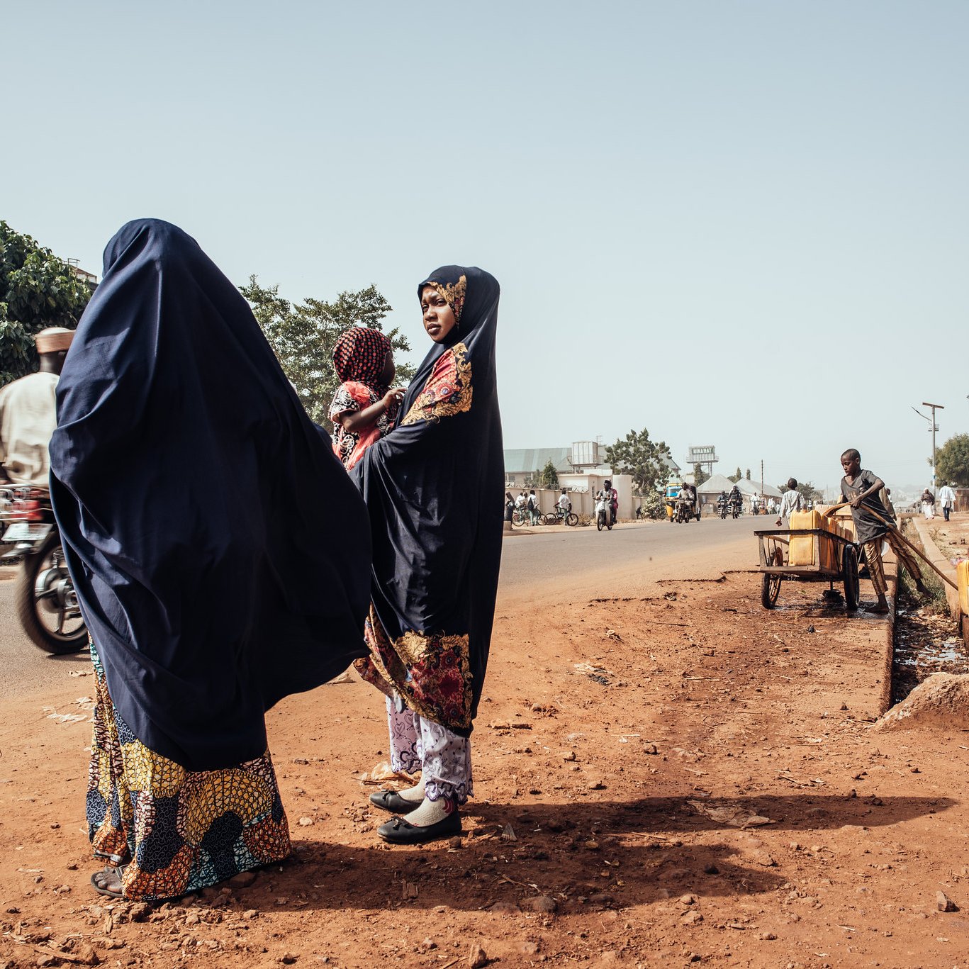 Dans cette image, nous sommes sur une route dans un environnement urbain. Sur le côté gauche, deux femmes portant des vêtements traditionnels de couleur sombre se tiennent ensemble. L'une d'elles a un sourire et semble discuter. Elles sont entourées de terre brune, et l'atmosphère est calme. Sur le côté droit, un homme est assis près d'un petit chariot, tandis qu'un panneau indique « GAGI », signalant un commerce local. On peut apercevoir d'autres personnes se déplaçant à pied ou à bicyclette sur la route, soulignant la vie active de ce lieu. Au fond, des arbres verdoyants ajoutent une touche de nature à l'environnement urbain.