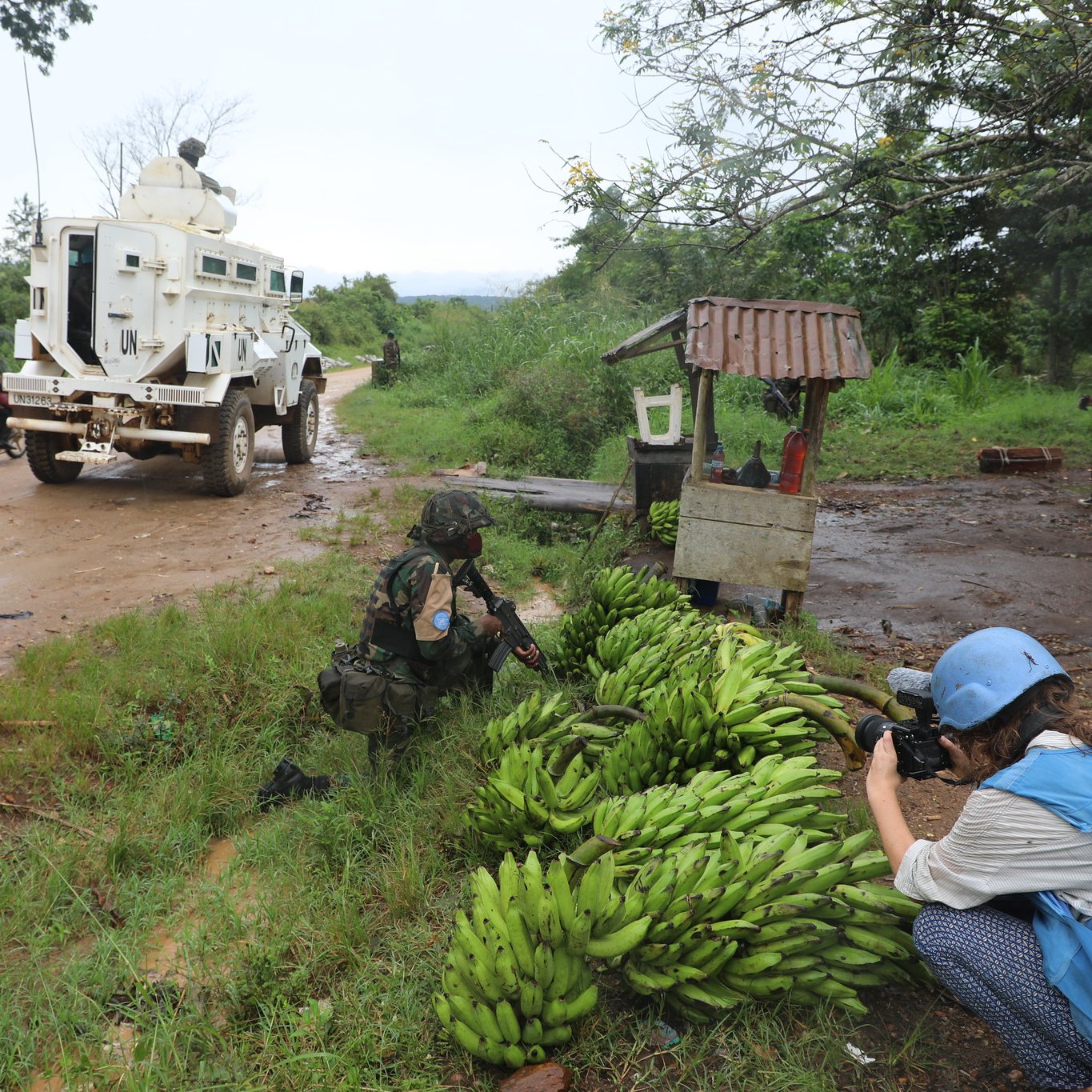L'image dépeint une scène rurale dans un environnement naturel. Sur le côté gauche, on aperçoit un groupe de bananes bien rangées au sol, d'un vert vibrant. À proximité, un soldat en uniforme utilise une caméra pour capturer des images, se penchant légèrement vers les bananes. Sur la droite, un véhicule blindé est garé sur une route poussiéreuse, entouré d'une végétation luxuriante. Au loin, d'autres personnes, comme un homme sur une moto et une femme à pied, ajoutent une touche de vie à cette scène. Le ciel semble nuageux, suggérant un temps humide, et l'atmosphère générale évoque un mélange d'activités militaires et de vie quotidienne.