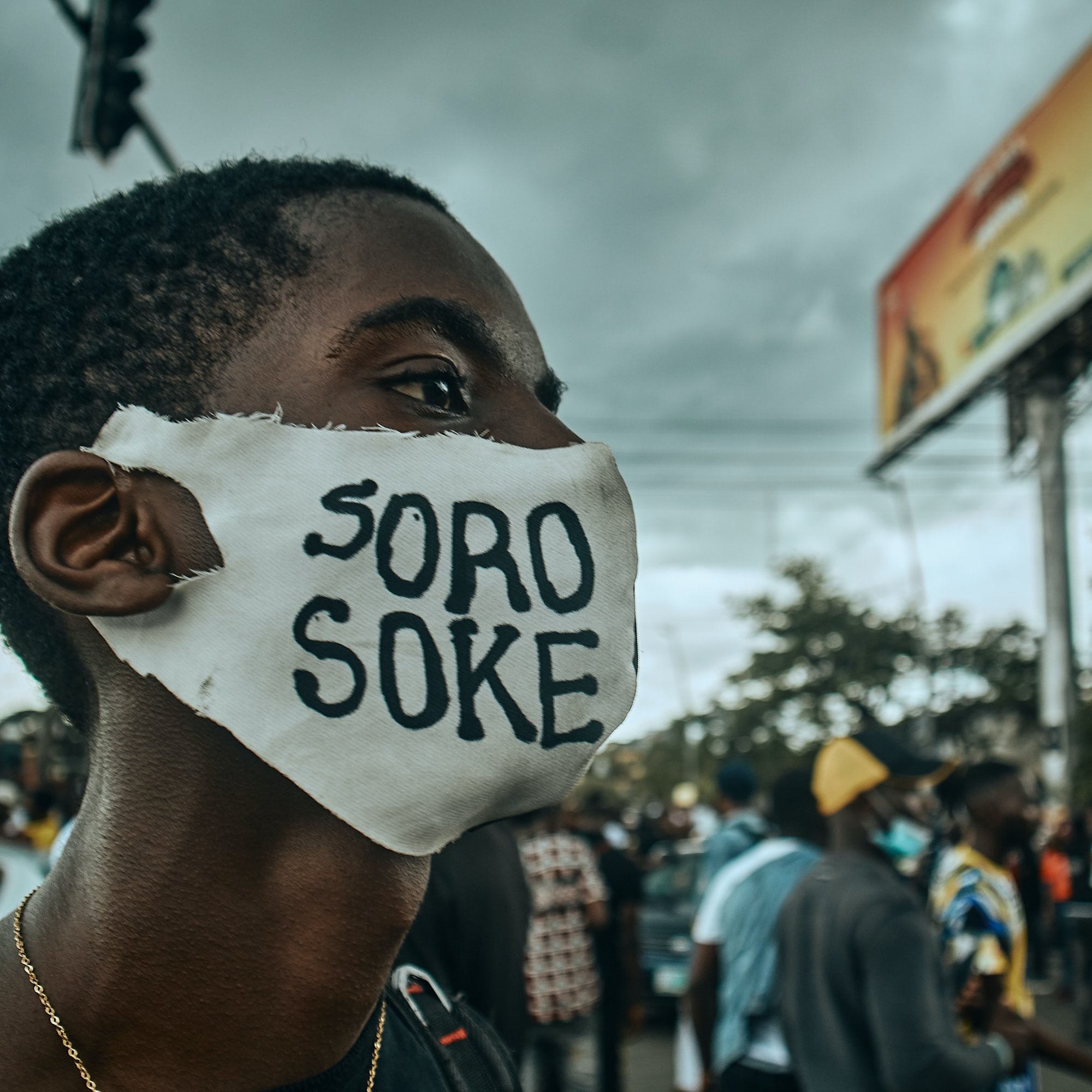 L'image montre une scène de manifestation dans une rue animée. Au premier plan, un jeune homme porte un masque blanc sur lequel est inscrit "SORO SOKE" en lettres noires, un slogan qui évoque la lutte pour les droits et la justice. Son regard est déterminé, et on peut sentir l'énergie de la foule qui l'entoure. À l'arrière-plan, des silhouettes de personnes se déplacent, et des affiches publicitaires peuvent être aperçues. Le ciel est nuageux, ce qui ajoute une atmosphère de tension à la scène. L'ensemble dégage une forte impression d'engagement et de solidarité.