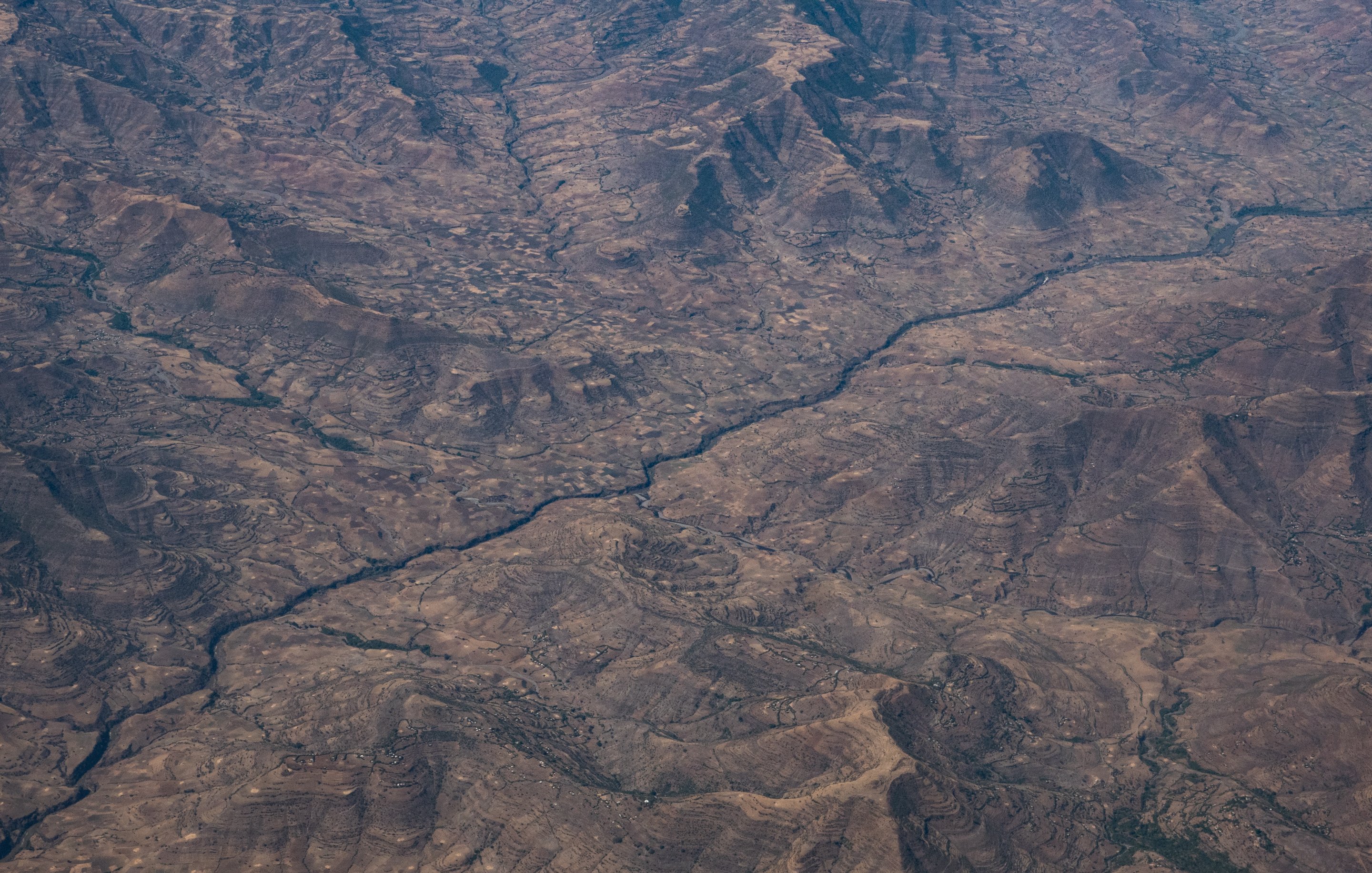 Cette image montre un paysage désertique vu du ciel. On peut observer de vastes étendues de terrain aride, striées par des vallées et des collines aux formes irrégulières. Au centre, un cours d'eau serpente à travers ce paysage, créant un contraste avec les zones sèches environnantes. Les couleurs dominantes sont des teintes de marron et de beige, évoquant un sol rocailleux et dénudé, tandis que la rivière apporte une touche de reflets plus sombres, suggérant des zones humides dans ce milieu. L'ensemble dégage une impression de vastitude et de sérénité, avec une nature sauvage encore peu altérée par l'homme.