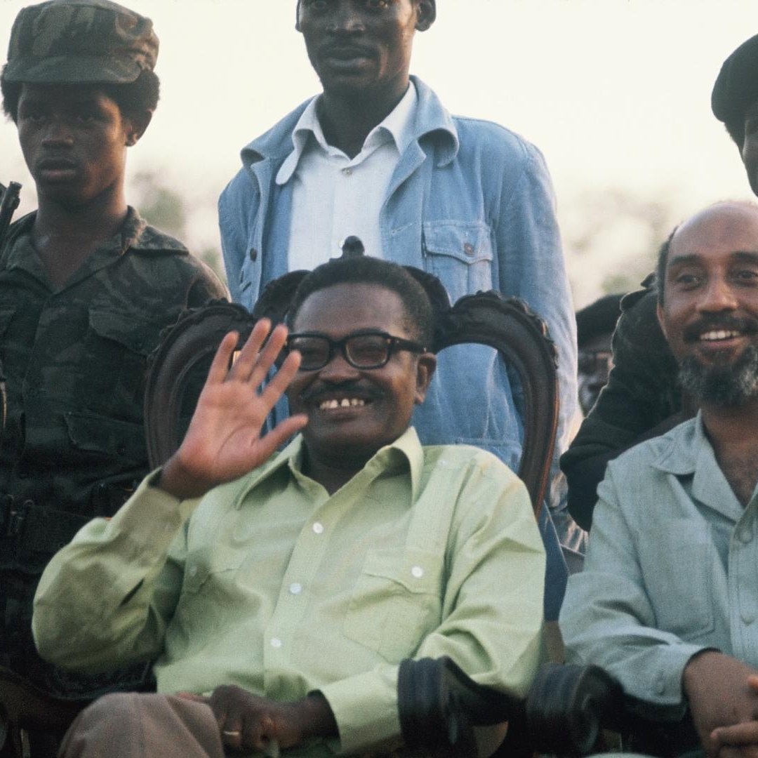 L'image présente un groupe de personnes, dont deux hommes au premier plan, assis sur des chaises ornées. L'un des hommes, souriant et portant des lunettes, salue de la main. Il a une chemise verte claire. À sa droite, un autre homme avec une chemise beige sourit également. En arrière-plan, plusieurs soldats en uniforme de camouflage et avec des casquettes se tiennent debout. L'environnement semble être en plein air, peut-être lors d'un événement officiel, avec un ciel illuminé par le crépuscule.