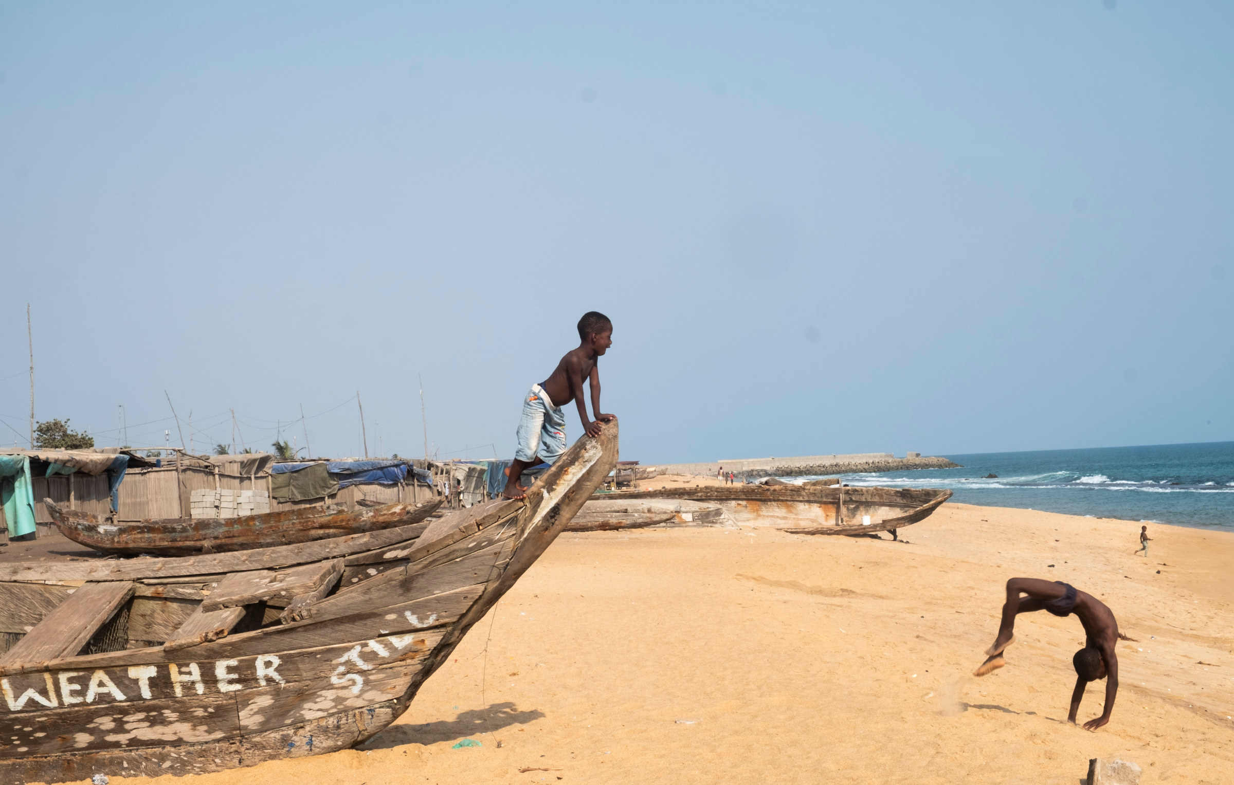 Dans cette scène vibrante, une femme porte une grande bassine sur sa tête, symbole de son travail quotidien. Elle avance avec assurance sur un chemin, tout en étant entourée de maisons à architecture simple, en bois et en bambou. Le ciel est clair, laissant présager une belle journée ensoleillée. À proximité, des enfants jouent ensemble, ajoutant une atmosphère de vie et de dynamisme à l'environnement. Les ombres et les textures des matériaux environnants se mêlent, créant un paysage riche et vivant. L'ensemble dégage une ambiance d'activité et de communauté.