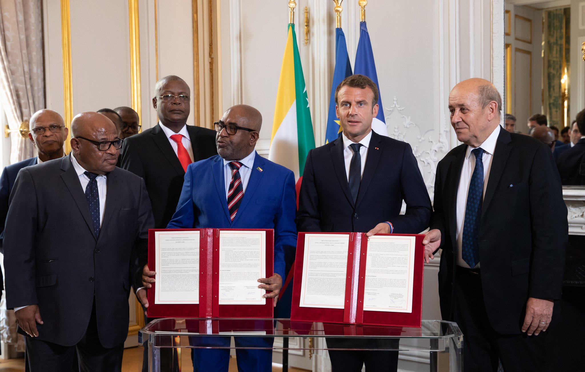 L'image montre un groupe de personnes lors d'une cérémonie officielle. Au centre, deux hommes tiennent des documents signés, symbolisant un accord ou un partenariat. L'un d'eux porte un costume bleu avec une cravate à motifs, tandis que l'autre est vêtu d'un costume sombre avec une cravate noir. En arrière-plan, on remarque des drapeaux, dont ceux de la France et d'autres pays africains. Les autres personnes en arrière-plan observant la scène portent également des costumes. L'ambiance générale est solennelle, mettant en avant un moment important de collaboration entre les nations.