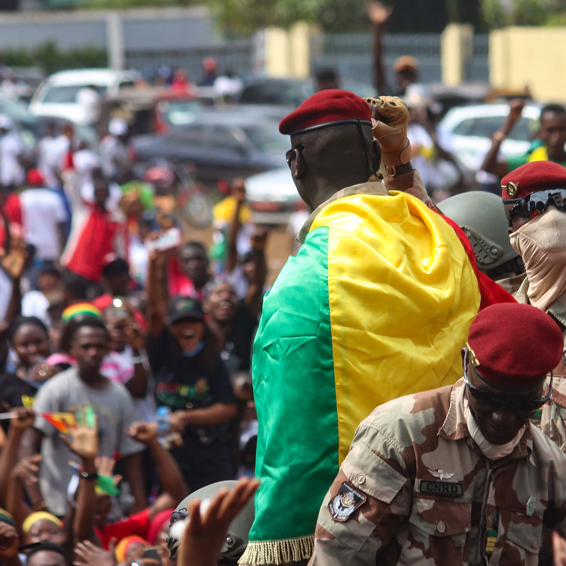 L'image montre une scène dynamique et pleine d'énergie. Au premier plan, un homme en uniforme militaire, portant un béret rouge, se tient sur une plateforme surélevée. Il est enveloppé dans un drapeau aux couleurs vives du Mali, qui est vert, jaune et rouge. Autour de lui, une foule enthousiaste s'agite, brandissant des drapeaux et exprimant leur joie. Les visages des personnes dans la foule montrent des émotions variées allant de l'excitation à la fierté. En arrière-plan, on peut voir des voitures stationnées et d'autres participants se mêlant dans une atmosphère de célébration collective. L'ensemble dégage un fort sentiment d'unité et de patriotisme.