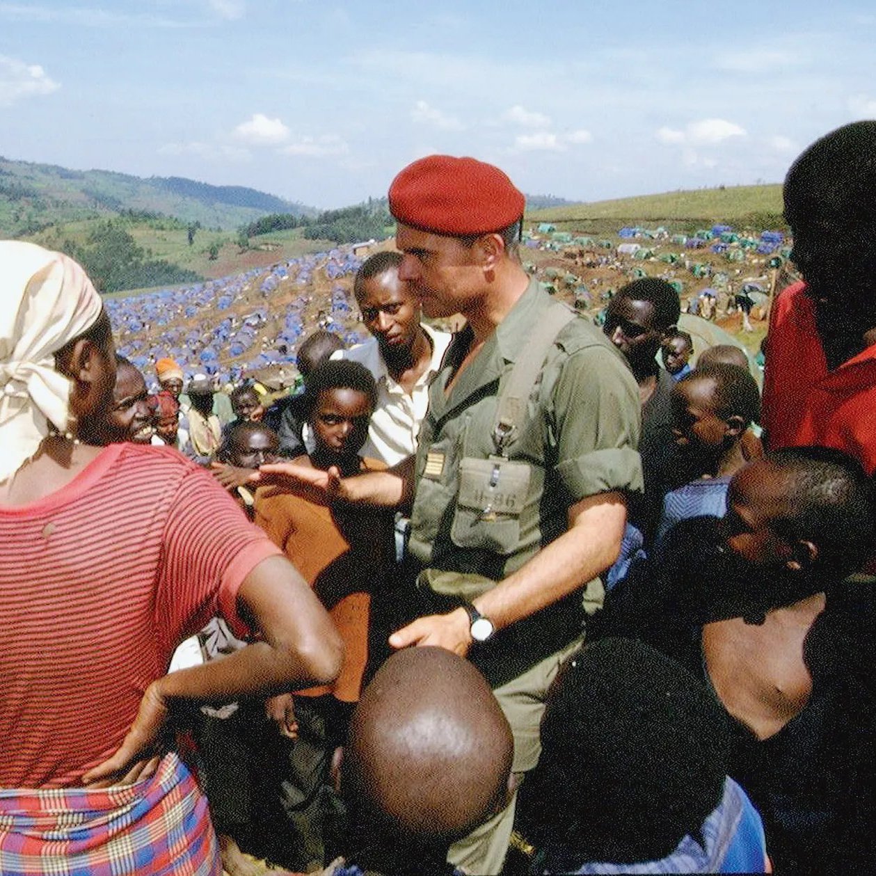 L'image montre un groupe de personnes dans un paysage ouvert et verdoyant. Au centre, un homme en uniforme militaire, portant un béret rouge, discute avec une femme qui porte un foulard et une blouse, le regard tourné vers lui. Elle semble attentive aux explications qu'il donne. Autour d'eux, plusieurs enfants et adultes, principalement d'origine africaine, écoutent avec intérêt. Les visages sont expressifs, certains avec des regards curieux. À l'arrière-plan, on aperçoit une vaste étendue de tentes bleues, indiquant probablement un camp de réfugiés ou de soutien. Le ciel est clair, avec quelques nuages, et les collines verdoyantes offrent une vue panoramique sur la scène. L'atmosphère dégage un sentiment de communauté et d'engagement.