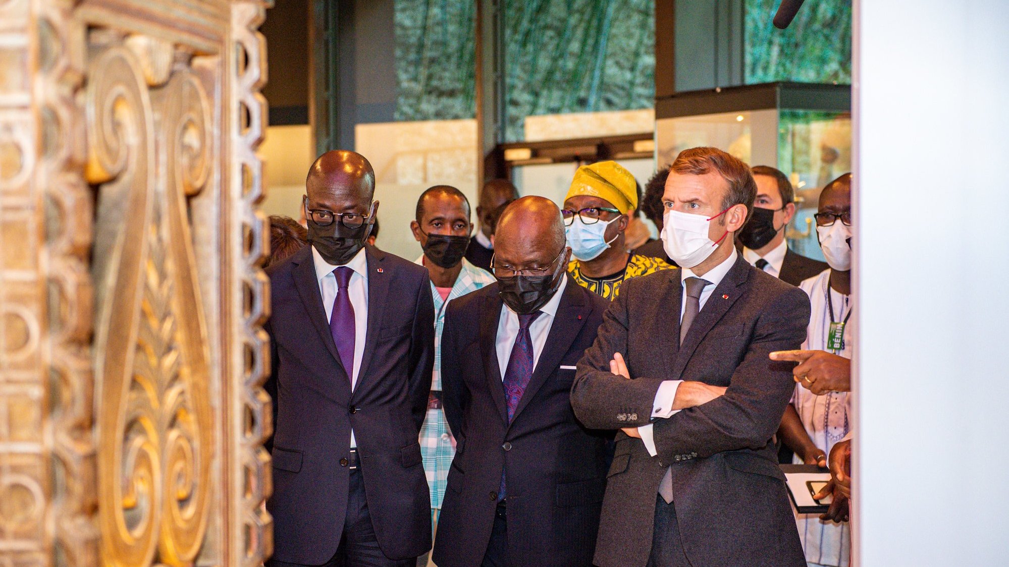 L'image montre un groupe de personnes, dont trois hommes, en train d'observer un objet d'art ou une exposition. Les hommes sont habillés de costumes formels, avec deux en costumes sombres et un en costume gris avec une cravate. Ils affichent des expressions de concentration et d'intérêt. À l'arrière-plan, on perçoit des éléments culturels, probablement des œuvres d'art ou des artifacts, créant une ambiance muséale. Les visiteurs portent tous des masques, ce qui indique qu'ils se trouvent dans un contexte où cela est nécessaire, peut-être en raison de préoccupations sanitaires. Le cadre est bien éclairé, mettant en valeur l'importance de l'exposition.