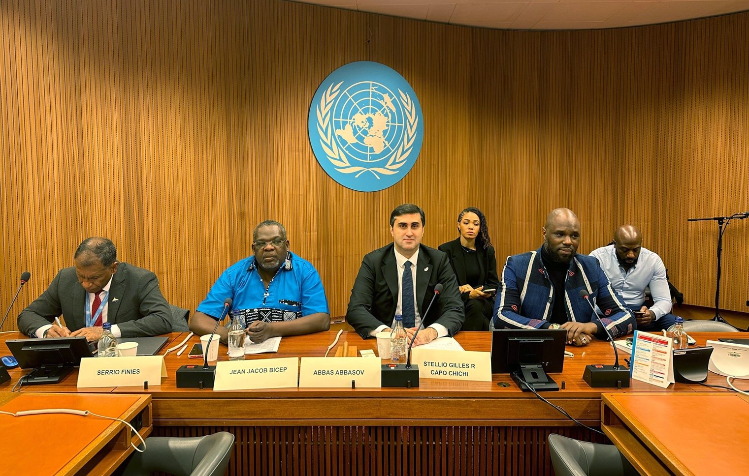 L'image montre une scène à une table de conférence, probablement lors d'une réunion aux Nations Unies. Au centre, un homme en costume noir et cravate blanche, avec un air sérieux, semble être le porte-parole ou le modérateur. À sa gauche, un homme portant une chemise bleue et des lunettes, avec un air attentif. À sa droite, un homme d'apparence plus décontractée avec un pull et une barbe, qui semble également participer à la discussion. En arrière-plan, plusieurs personnes sont assises, écoutant attentivement. Le décor est sobre, avec des murs en bois et le logo des Nations Unies en arrière-plan.