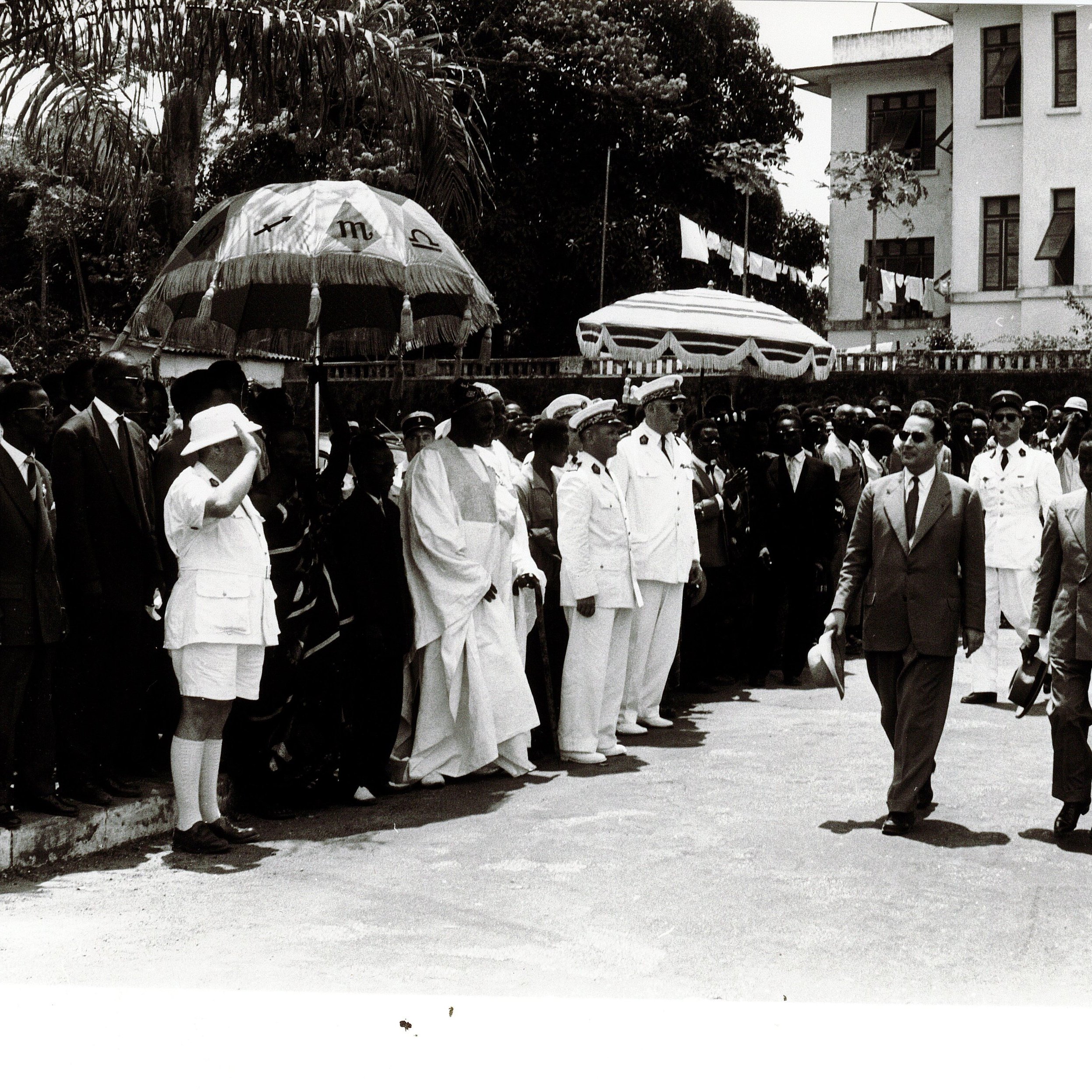 L'image montre un groupe de personnes rassemblées en extérieur, probablement lors d'un événement officiel. Au centre, des hommes en costumes formels marchent, entourés de dignitaires et de membres du public. Certains portent des uniformes militaires, tandis que d'autres sont habillés de manière civile. Il y a également des parapluies décoratifs en arrière-plan. L'atmosphère semble solennelle, et l'environnement suggère une atmosphère tropicale avec des arbres en arrière-plan. La scène donne une impression de cérémonie et de célébration.