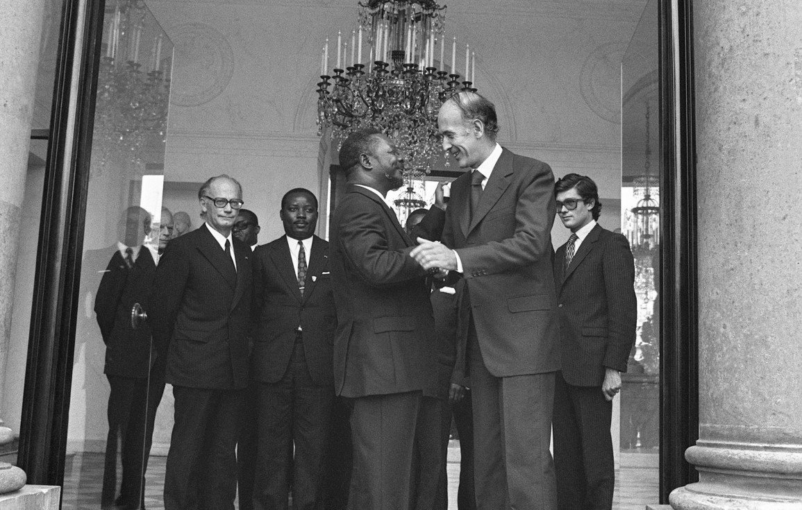 L'image montre un groupe d'hommes en costume, réunis dans un grand hall bien éclairé. Au centre, deux hommes se serrent la main, affichant des expressions de cordialité. L'un est légèrement plus âgé, avec des cheveux grisonnants, tandis que l'autre est plus jeune. En arrière-plan, plusieurs autres hommes les observent, tous vêtus de costumes sombres, ce qui accentue l'atmosphère formelle de la rencontre. Le décor est élégant, avec un grand lustre suspendu au plafond et des colonnes en pierre qui ajoutent une touche de sophistication.