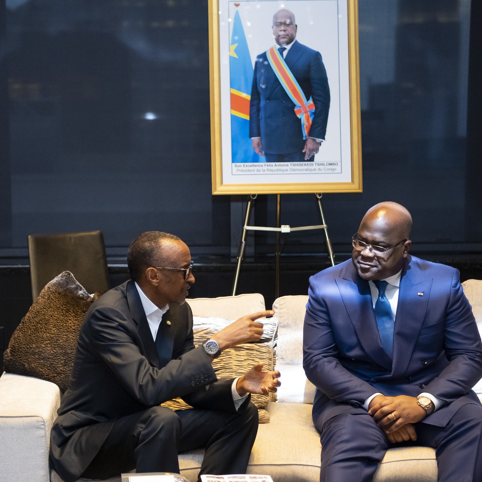 L'image montre deux hommes assis sur un canapé dans un cadre intérieur élégant. L'un des hommes porte un costume noir et semble s'engager dans une conversation. L'autre homme est habillé d'un costume bleu et sourit. En arrière-plan, il y a un portrait encadré d'un homme en costume, avec un drapeau de la République démocratique du Congo visible derrière lui. L'environnement est moderne, avec des luminaires doux et un décor soigné, suggérant une ambiance formelle ou diplomatique.