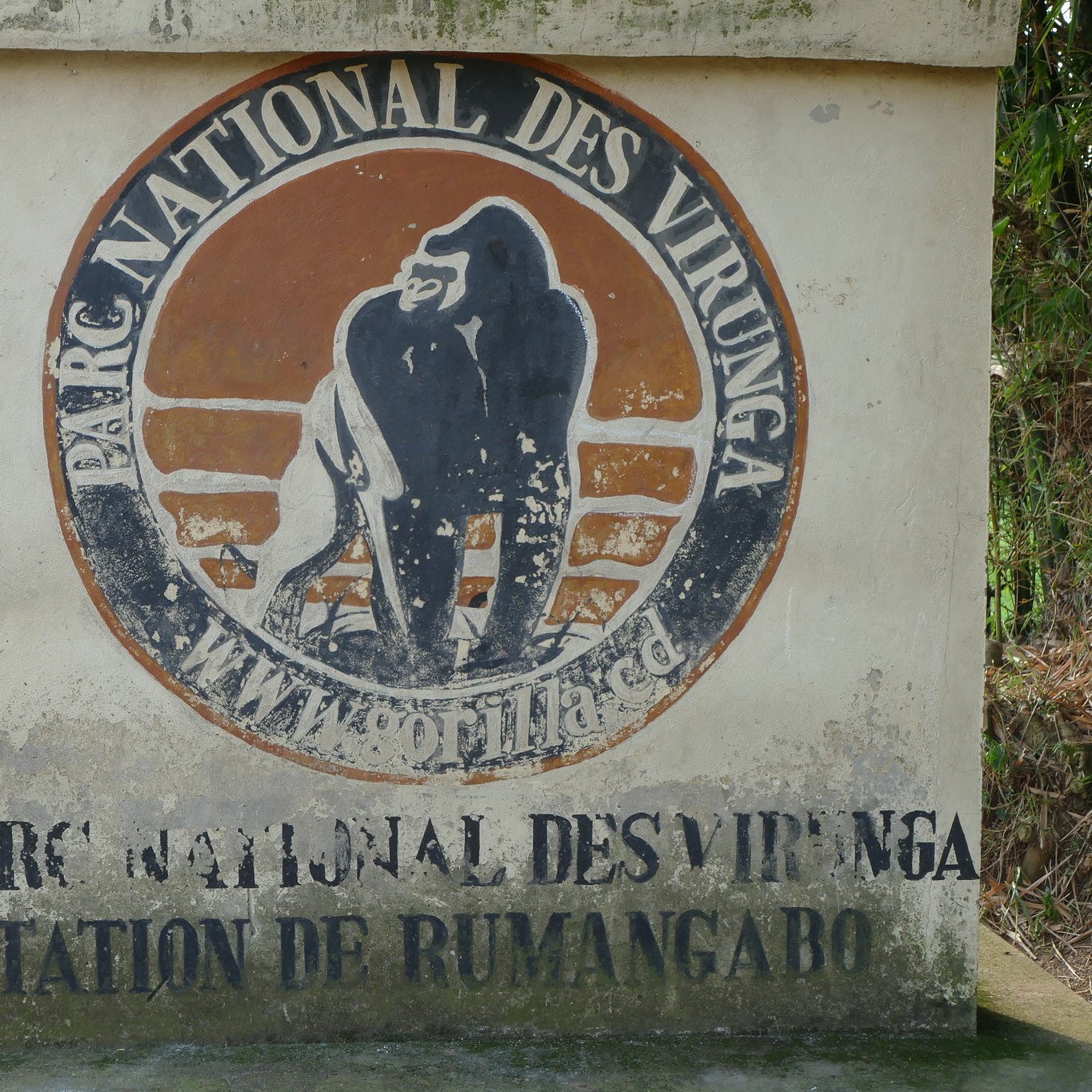 The image features a sign marking the entrance to the Virunga National Park. The sign displays the park's name in both French and English, showcasing the emblem of the park, which includes an illustration of a gorilla. The surroundings seem to depict a rural area, with a road leading away from the entrance and a building visible in the background. The overall atmosphere suggests an area of natural beauty and wildlife conservation.