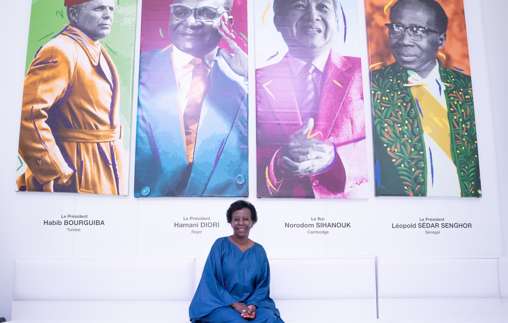 L'image présente une femme assise sur un banc dans une galerie moderne. Derrière elle, il y a quatre portraits colorés de figures politiques africaines. Chaque portrait est visiblement distinct, avec des couleurs vives, représentant des hommes portant des costumes traditionnels ou modernes. La femme, vêtue d'une robe bleu foncé, sourit légèrement et semble apprécier l'exposition. Le décor est minimaliste, avec des sièges blancs et des poufs de différentes couleurs qui ajoutent une touche de dynamisme à l'ensemble. L'ambiance générale est accueillante et artistique, invitant à la réflexion sur l'histoire politique de l'Afrique.