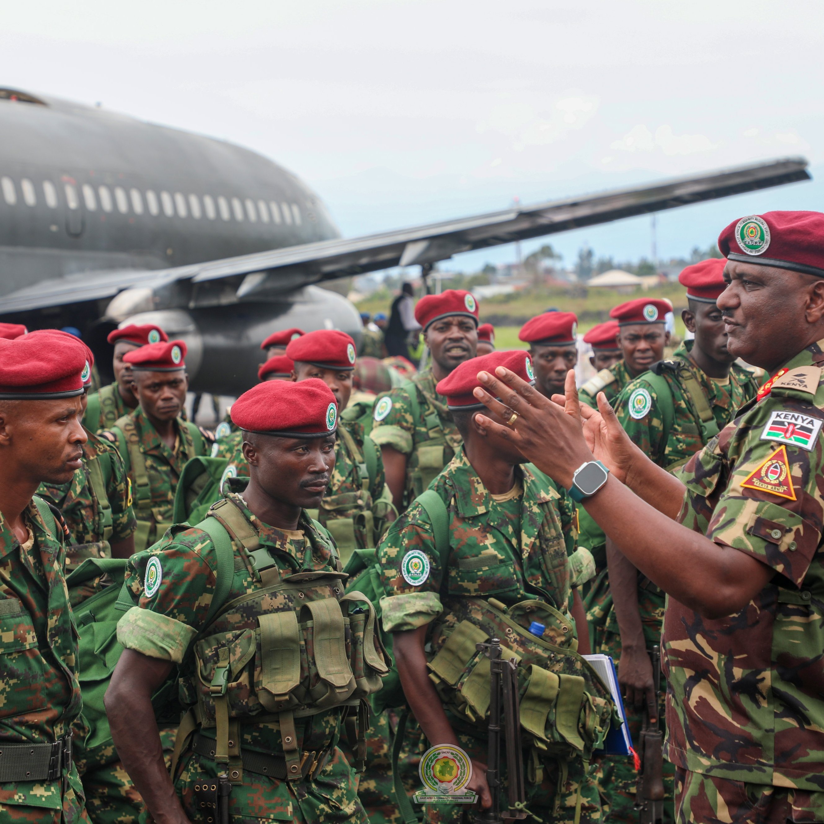 L'image montre un groupe de soldats en uniforme militaire, réunis près d'un gros avion de transport. Les soldats portent des casques et des berets rouges, et ils sont habillés de camouflages verts. L'un des officiers, qui semble donner des instructions ou des encouragements, se tient au centre, gesticulant avec les mains. En arrière-plan, on peut voir l'avion de taille importante, avec des couleurs de camouflage, sur un terrain d'aviation, tandis que le ciel est légèrement nuageux. L'atmosphère est sérieuse, témoignant d'une mobilisation militaire.