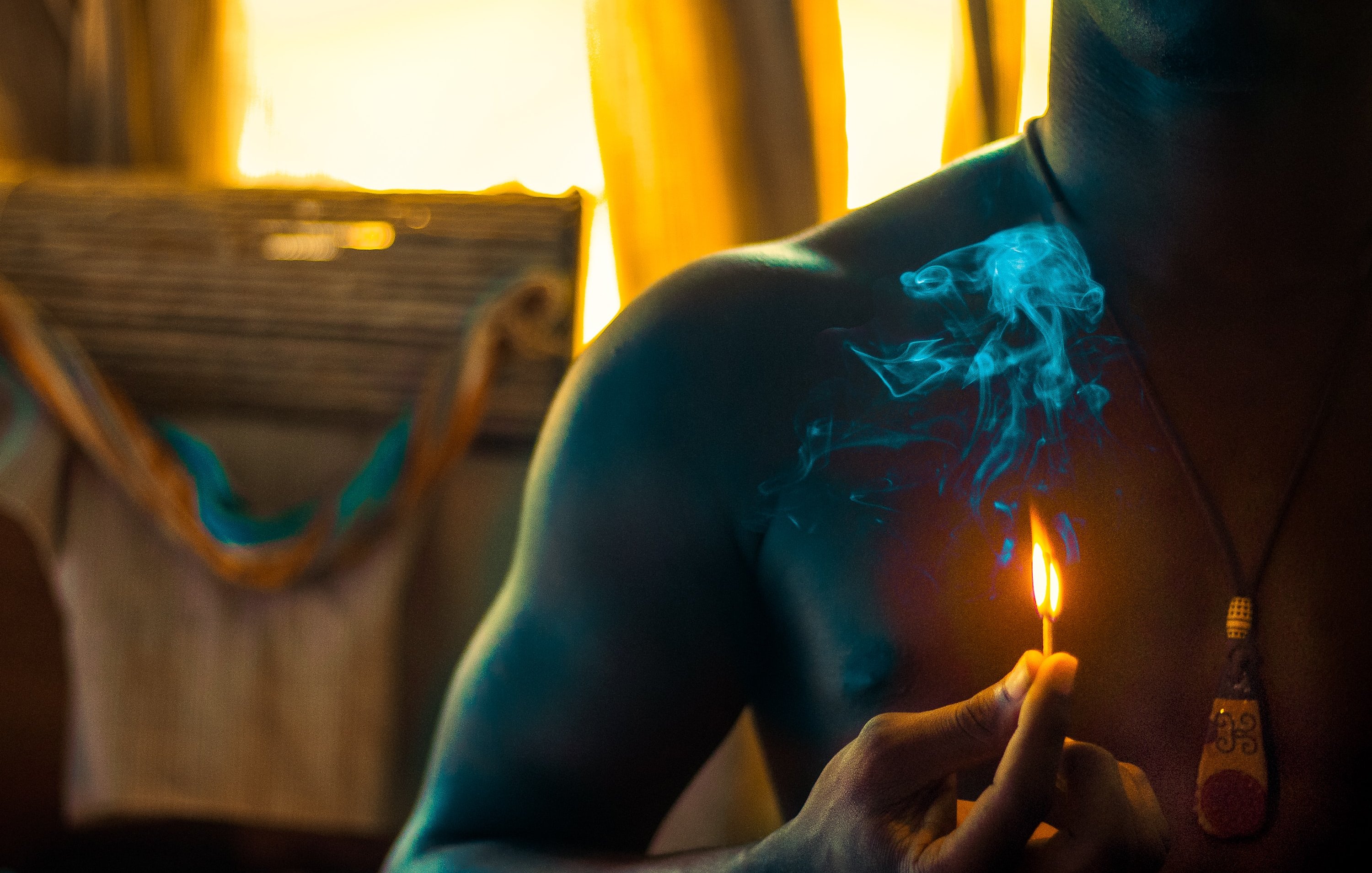 Dans cette image, nous voyons un homme torse nu tenant une petite flamme d'une allumette qui fume légèrement. La lumière douce qui l'entoure crée une ambiance chaleureuse. À l'arrière-plan, des éléments flous suggèrent un environnement intérieur, peut-être une pièce éclairée par le soleil couchant, ce qui ajoute une touche de chaleur à l'atmosphère. La fumée s'élève lentement, créant un contrasté avec la lueur de la flamme.