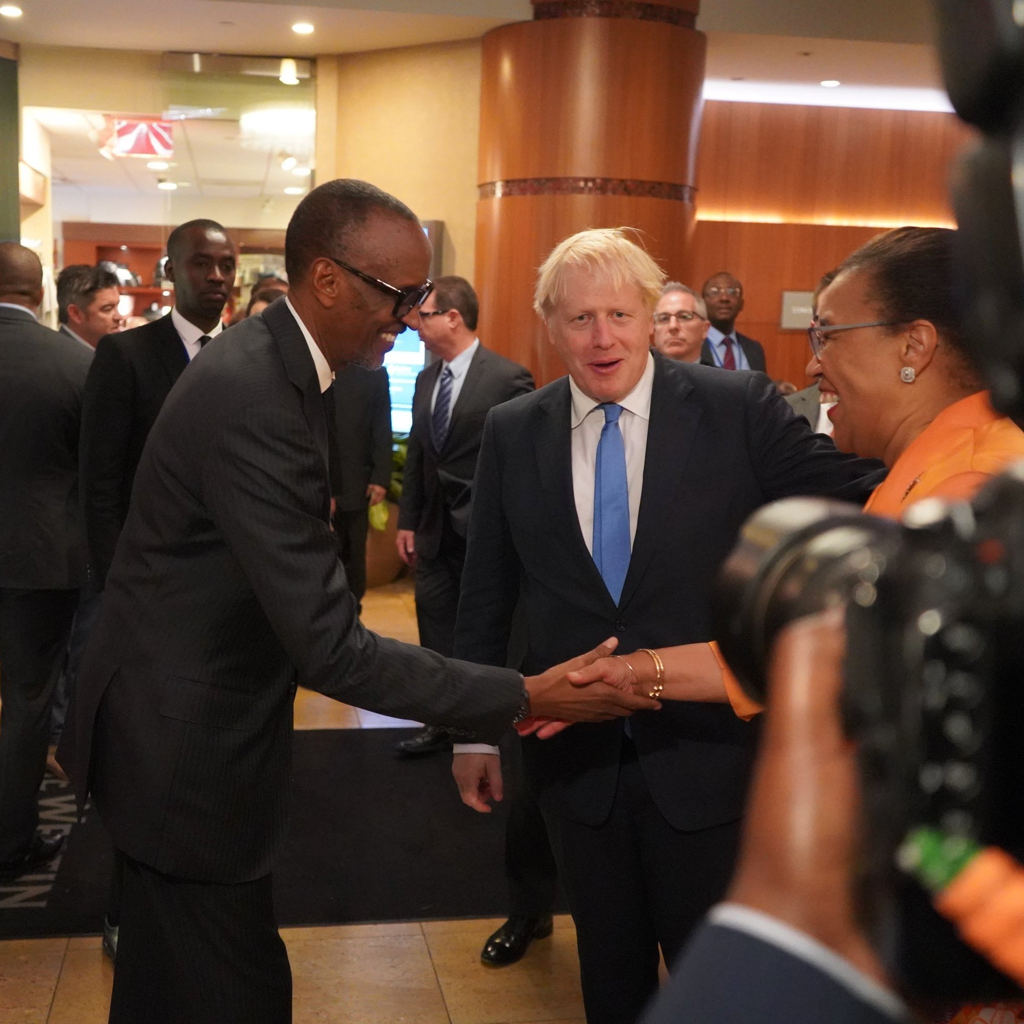 L'image représente une scène dans un hall d'hôtel ou de conférence, où plusieurs personnes sont rassemblées. Au centre, deux hommes se serrent la main : l'un est de grande taille, portant un costume sombre et des lunettes, tandis que l'autre, avec des cheveux blonds, est habillé d'un costume clair. Ils affichent tous deux des sourires amicaux. En arrière-plan, on peut voir plusieurs autres personnes en discussion, ainsi que des détails du décor intérieur, comme des murs aux tons chauds et des éléments de lumière. L'ambiance semble professionnelle et conviviale.