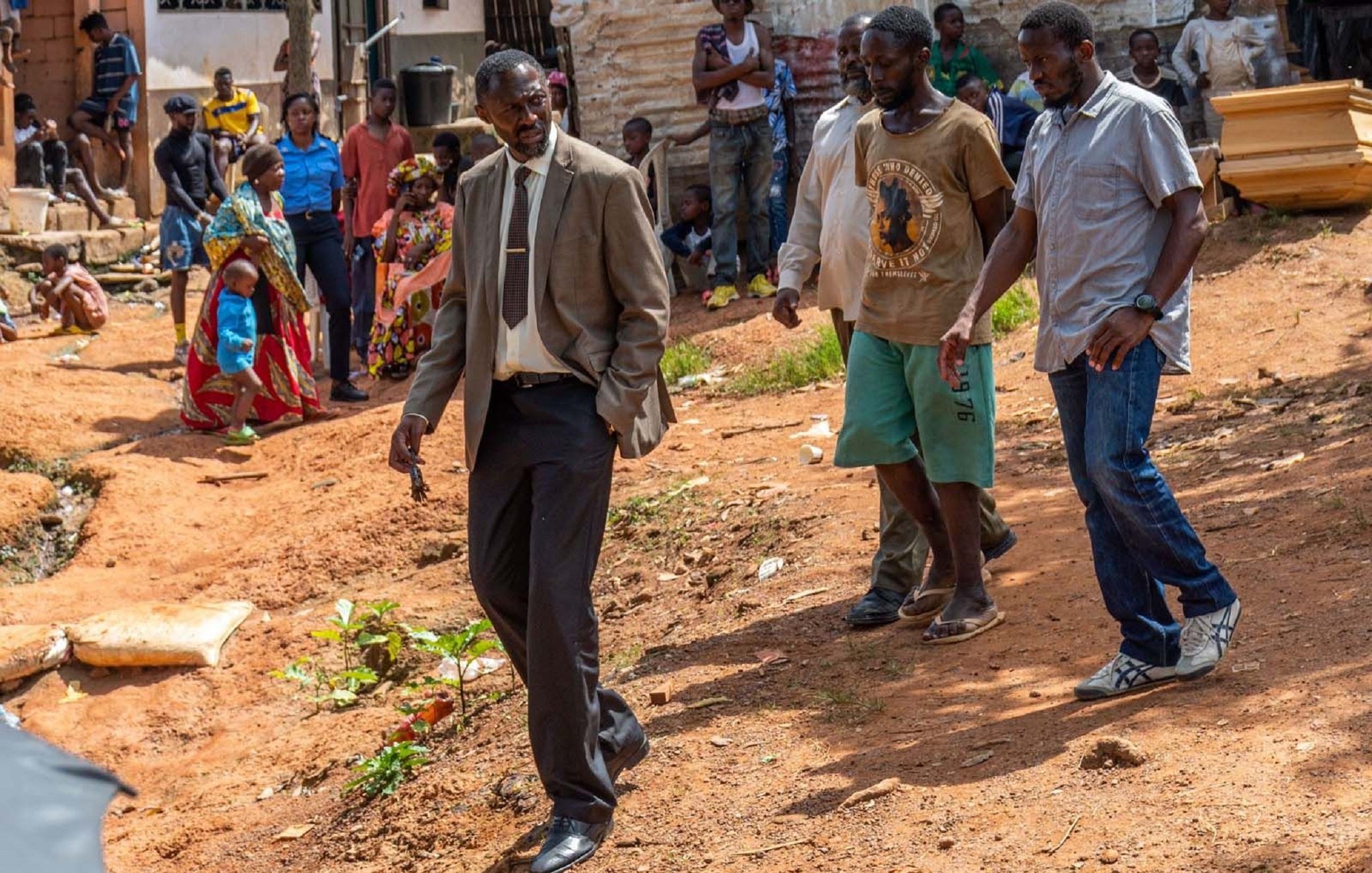 L'image montre un groupe de personnes dans un environnement rural ou semi-urbain. Au premier plan, un homme en costume cravate semble marcher avec assurance, le regard concentré. À sa gauche, un homme portant un t-shirt et un short se tient à côté de lui. D'autres personnes, habillées de manière décontractée, se déplacent également dans la scène, certains sont occupés à interagir entre eux. Le sol est en terre battue et il y a quelques plantes éparses. En arrière-plan, on peut apercevoir des maisons et des groupes de personnes qui discutent. L'atmosphère semble conviviale mais déterminée, avec un sentiment de communauté visible parmi les participants.