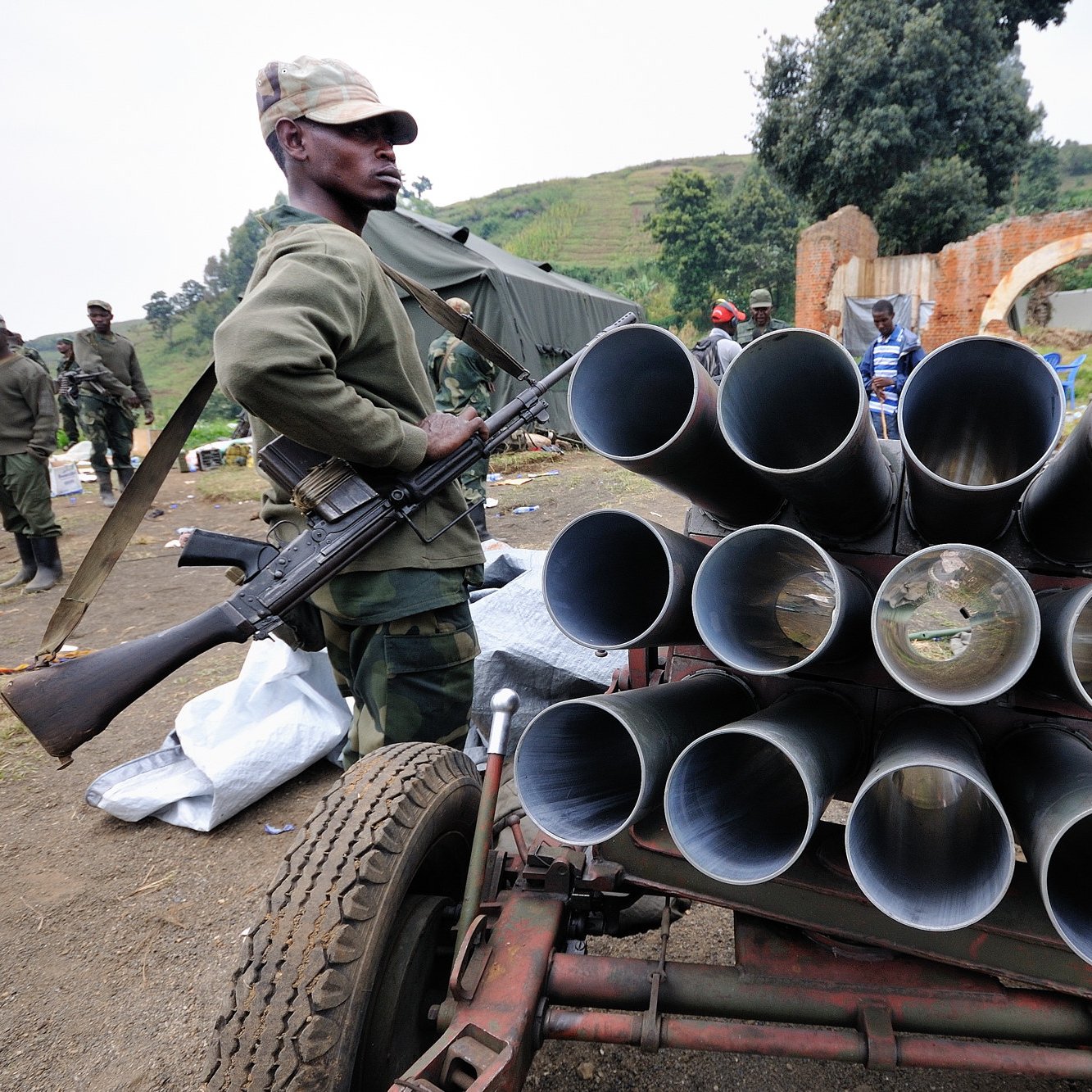 L'image montre un soldat en uniforme, debout à côté d'un véhicule équipé de plusieurs tubes visibles en rangée, qui semblent être des lance-roquettes. Le soldat tient une arme à feu, se tenant avec sérieux. En arrière-plan, on aperçoit d'autres soldats, ainsi qu'un environnement chaotique où des objets sont éparpillés, ce qui donne l'impression d'une zone de conflit ou de préparation militaire. Le tout se déroule dans un paysage verdoyant, avec des collines au loin.