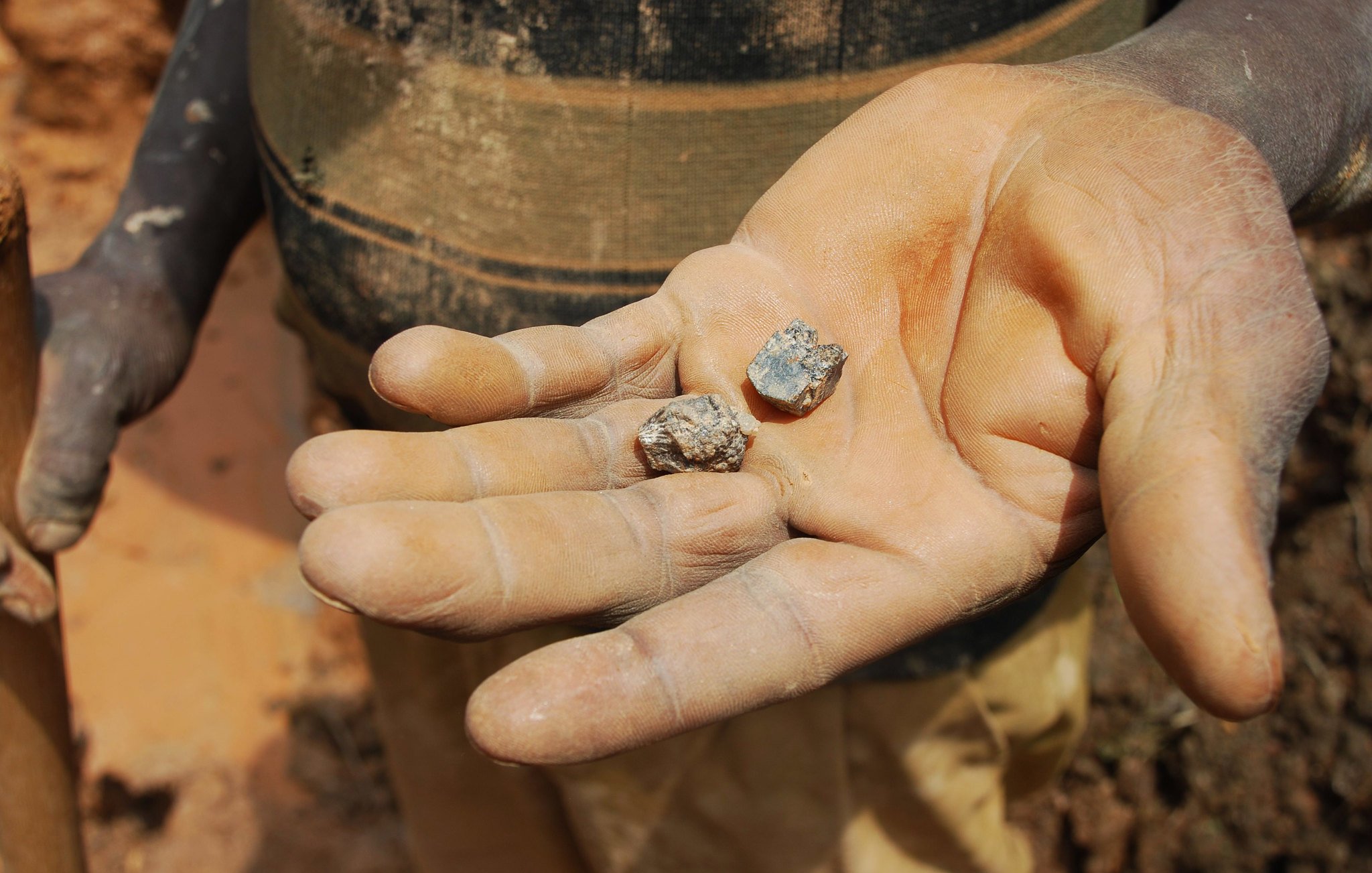 L'image montre une main, visiblement usée et sale, qui tient deux pierres. La paume est ouverte, révélant les deux morceaux de minéraux. L'un semble être plus gros et terne, tandis que l'autre est plus petit et a une surface légèrement brillante. Le fond de l'image montre un sol de terre battue, ce qui suggère que la scène se déroule dans un environnement minier ou rural. La lumière éclaire les pierres, mettant en avant leurs textures rugueuses et leurs formes irrégulières.