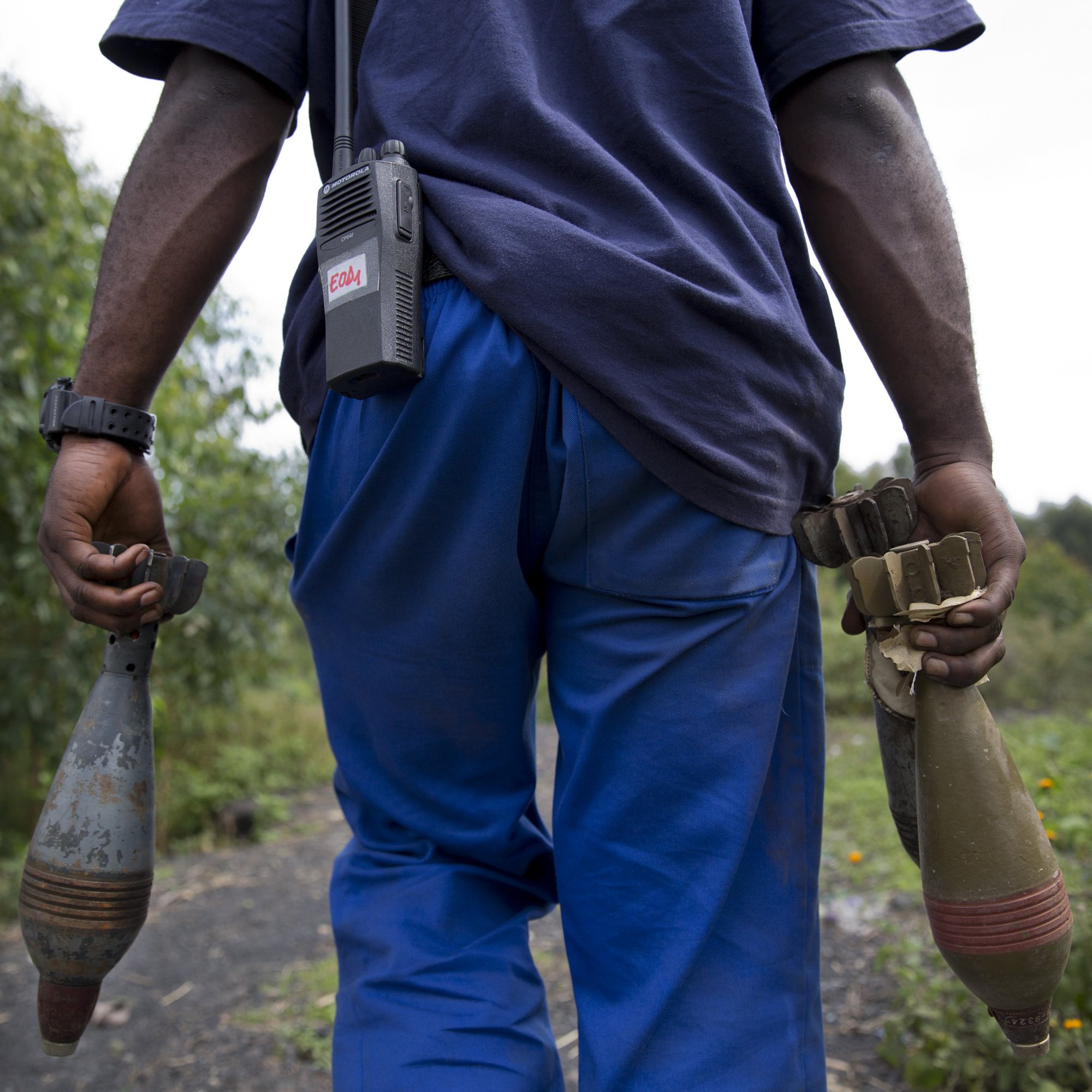 L'image montre un homme vu de dos, portant des vêtements décontractés : un t-shirt bleu foncé et un pantalon bleu. Il tient dans chaque main un objet long et cylindrique, probablement des munitions ou des dispositifs explosifs. À sa taille, il y a un appareil de communication. En arrière-plan, on aperçoit une nature verdoyante, avec des arbres et des plantes. L'ambiance semble à la fois sérieuse et chargée d'une importance qui nécessite prudence et vigilance.