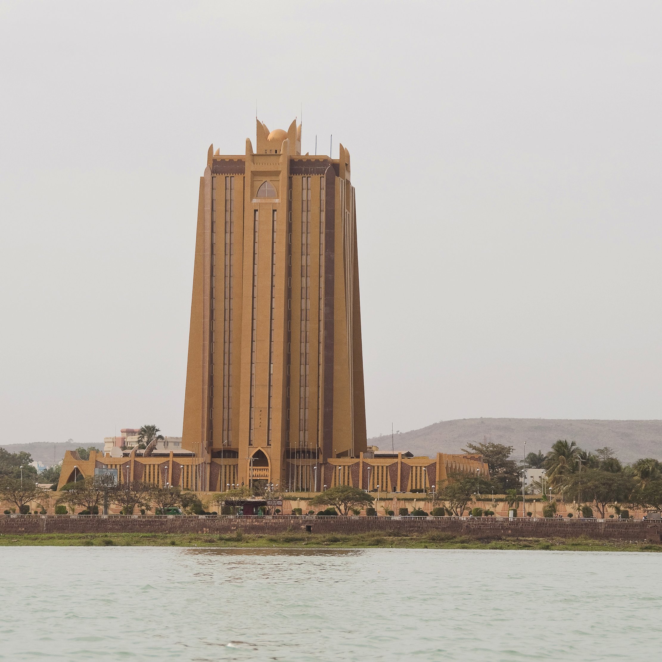L'image montre un bâtiment imposant qui semble être une tour, située près de l'eau. Ce bâtiment, d'une teinte dorée, a une architecture moderne et élancée. Sa structure se soulève vers le ciel, avec des lignes verticales marquées et des détails décoratifs au sommet, donnant une impression de grandeur. En arrière-plan, on aperçoit quelques collines et des arbres, tandis qu'au premier plan, on voit la surface calme de l'eau. Le ciel est légèrement nuageux, créant une ambiance tranquille et légèrement brumeuse autour de cette scène urbaine.