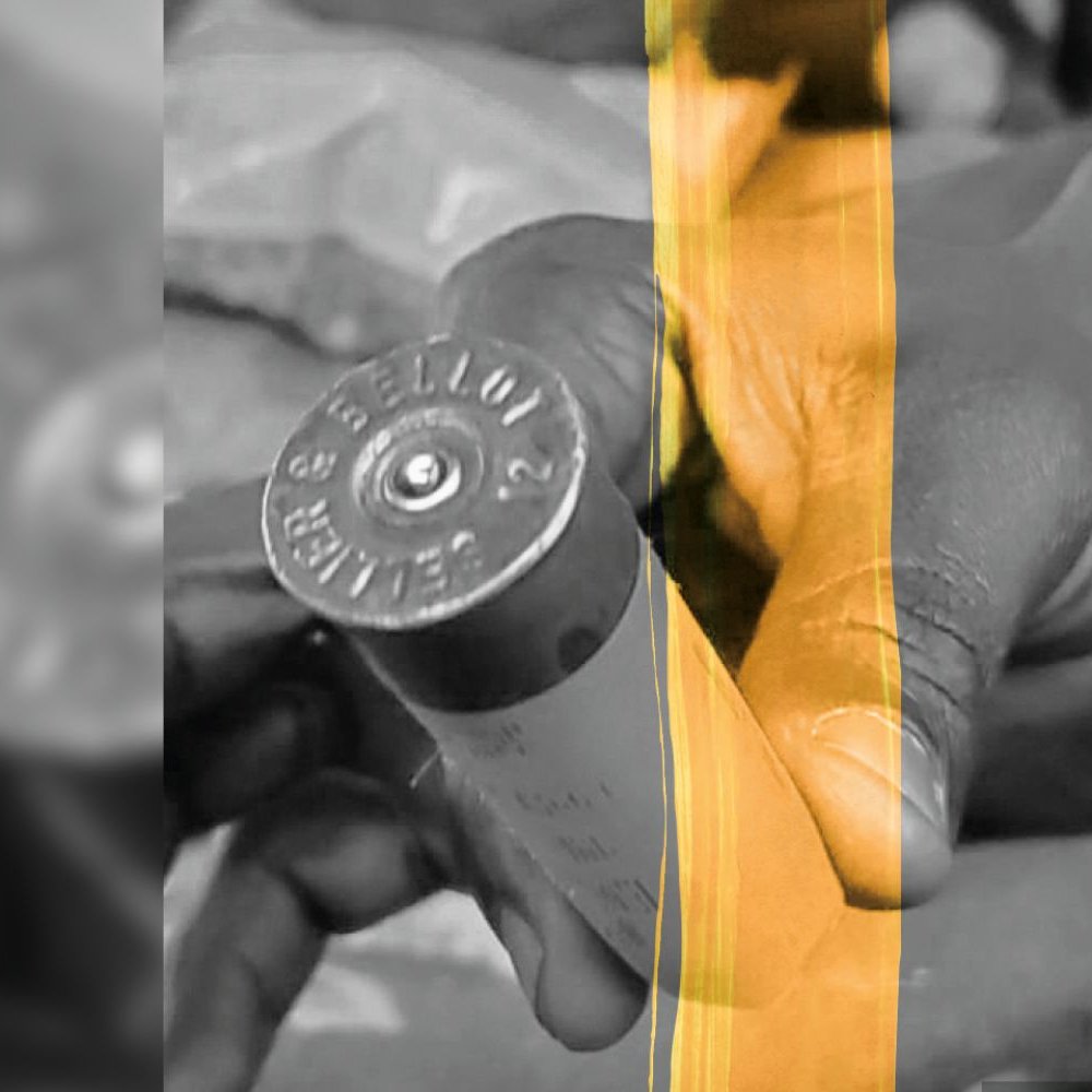 L'image montre deux mains tenant une cartouche de balle. La cartouche est de taille cylindrique et présente des inscriptions sur le côté, ainsi qu'un fond plat. Les mains sont légèrement enroulées autour de la cartouche, montrant un geste de manipulation. Le fond est flou, mettant en avant l'objet au centre de l'image. Les couleurs semblent être réduites à des nuances de noir et blanc, accentuant le contraste entre les mains et la cartouche. L'atmosphère générale est sérieuse, indiquant peut-être une situation de travail ou de préparation.