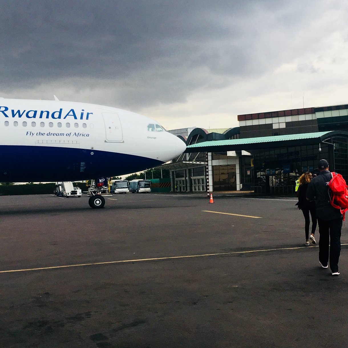 L'image montre un groupe de personnes marchant vers un avion de la compagnie RwandAir, qui est garé sur une aire d'aéroport. L'avion, avec une livrée bleue et blanche, est imposant avec un nez bien visible. Le ciel est nuageux, laissant présager une ambiance légèrement sombre. À l'arrière-plan, on voit un bâtiment d'aéroport avec une architecture moderne. Les voyageurs, portant des sacs à dos et d'autres bagages, avancent vers l'avion, créant un sentiment de départ et d'aventure. Le sol est en béton, et il y a des cônes de signalisation dispersés sur le terrain.