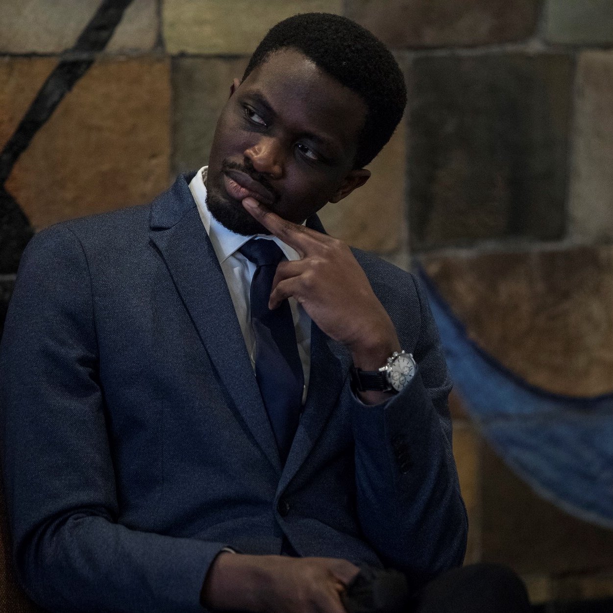 L'image montre un homme assis dans un environnement intérieur. Il porte un costume sombre avec une chemise claire et une cravate, ce qui dénote un certain formalisme. Son expression est pensive, avec un doigt sur le menton, suggérant qu'il réfléchit à quelque chose de sérieux. En arrière-plan, on aperçoit un mur aux motifs colorés, avec des formes géométriques qui ajoutent une touche artistique à la scène. L'ambiance est calme et réfléchie.