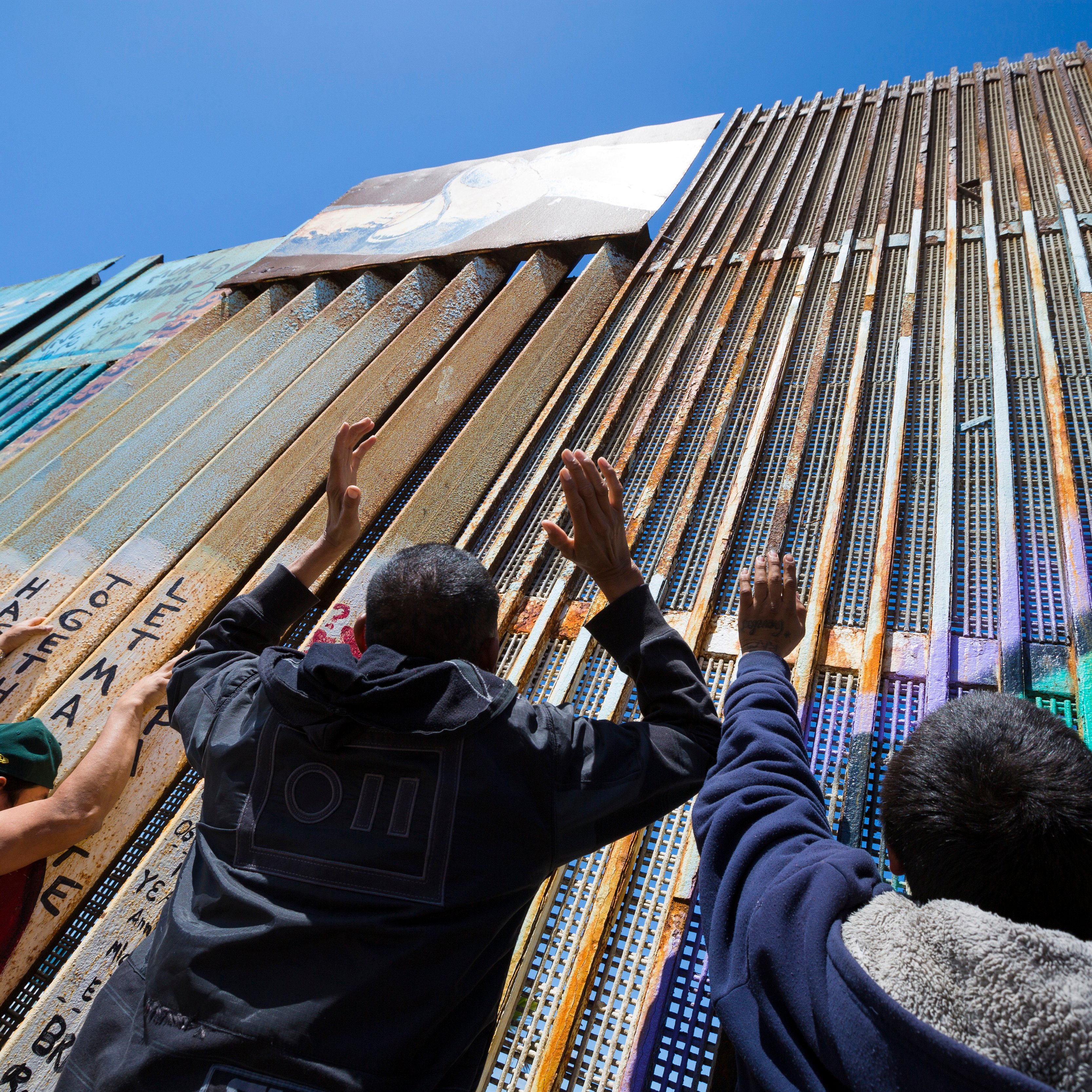 Sur cette image, on peut voir un grand mur en métal qui sépare deux zones. Le mur est composé de panneaux de différentes textures et couleurs, certains recouverts de graffitis. Au premier plan, on aperçoit plusieurs personnes qui tendent les mains vers le mur, comme si elles cherchaient un contact ou un lien. Le ciel est bleu et dégagé en arrière-plan, ce qui crée un contraste avec le métal de la barrière. L'atmosphère semble chargée d'émotion et de désir de connexion.