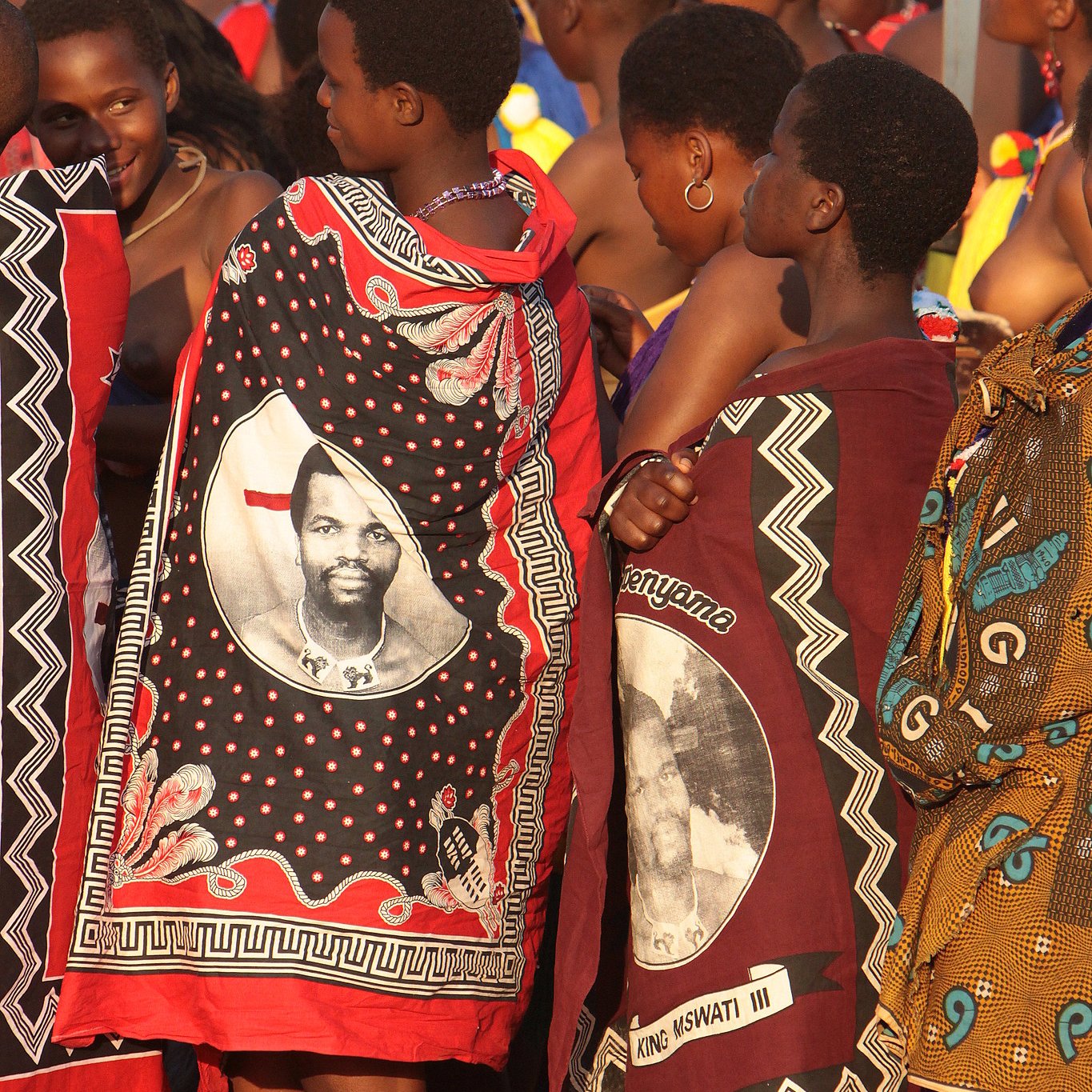 Dans cette image, on voit un groupe de femmes habillées de façon traditionnelle, revêtues de vêtements colorés et ornés de motifs riches. Certaines portent des châles qui affichent des portraits d'un homme, probablement une figure importante de leur culture. Les femmes se tiennent proches les unes des autres, exprimant un esprit de solidarité. L'atmosphère est vibrante, suggérant une célébration ou un événement culturel, avec des couleurs vives qui évoquent une culture riche et dynamique. Les sons environnants pourraient comprendre des musiques folkloriques et des rires, ajoutant à l'ambiance festive.